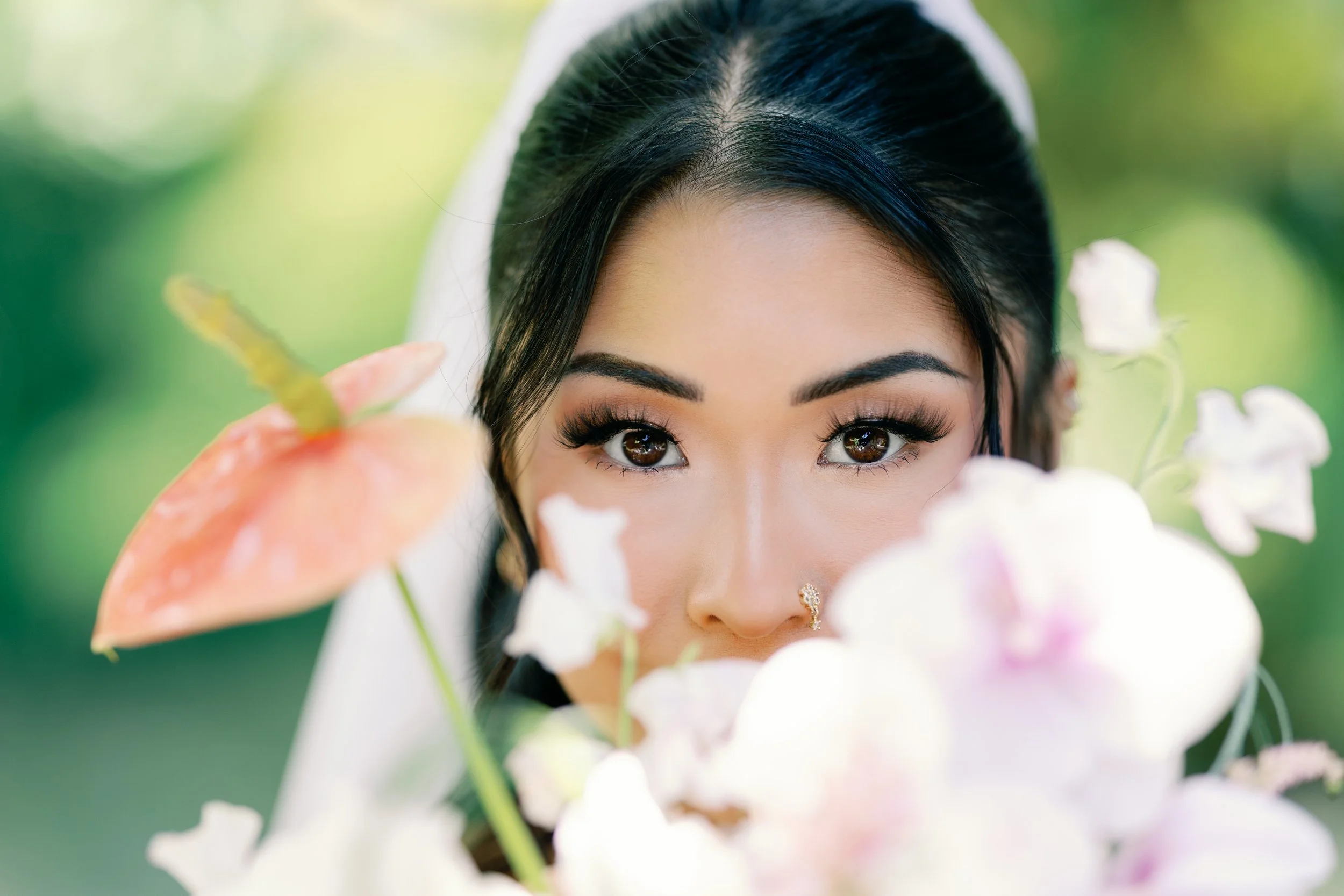 A bride with dark hair and makeup peeking through white flowers and green foliage outdoors.