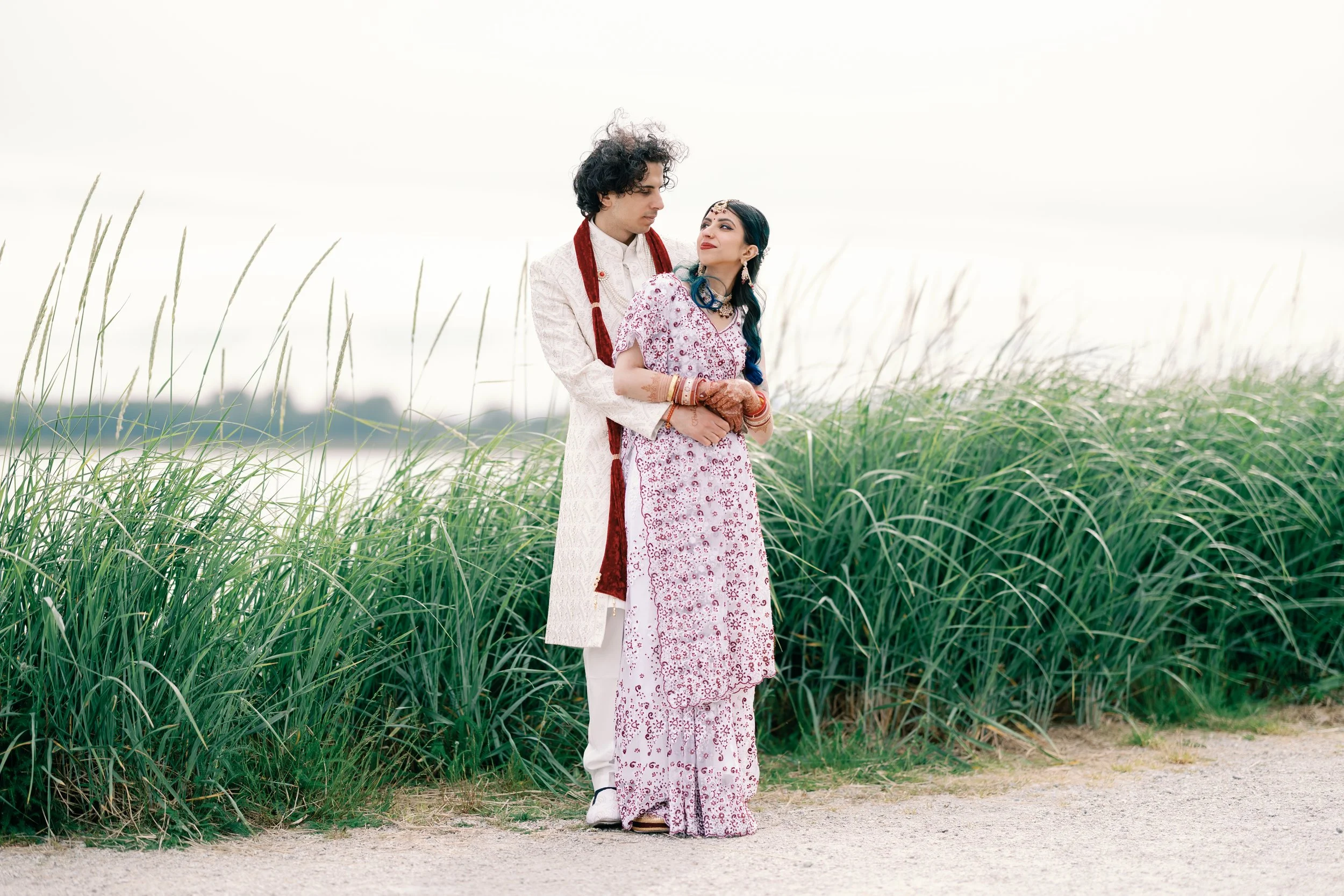 A wedding couple dressed in traditional Indian attire standing in a grassy outdoor setting.