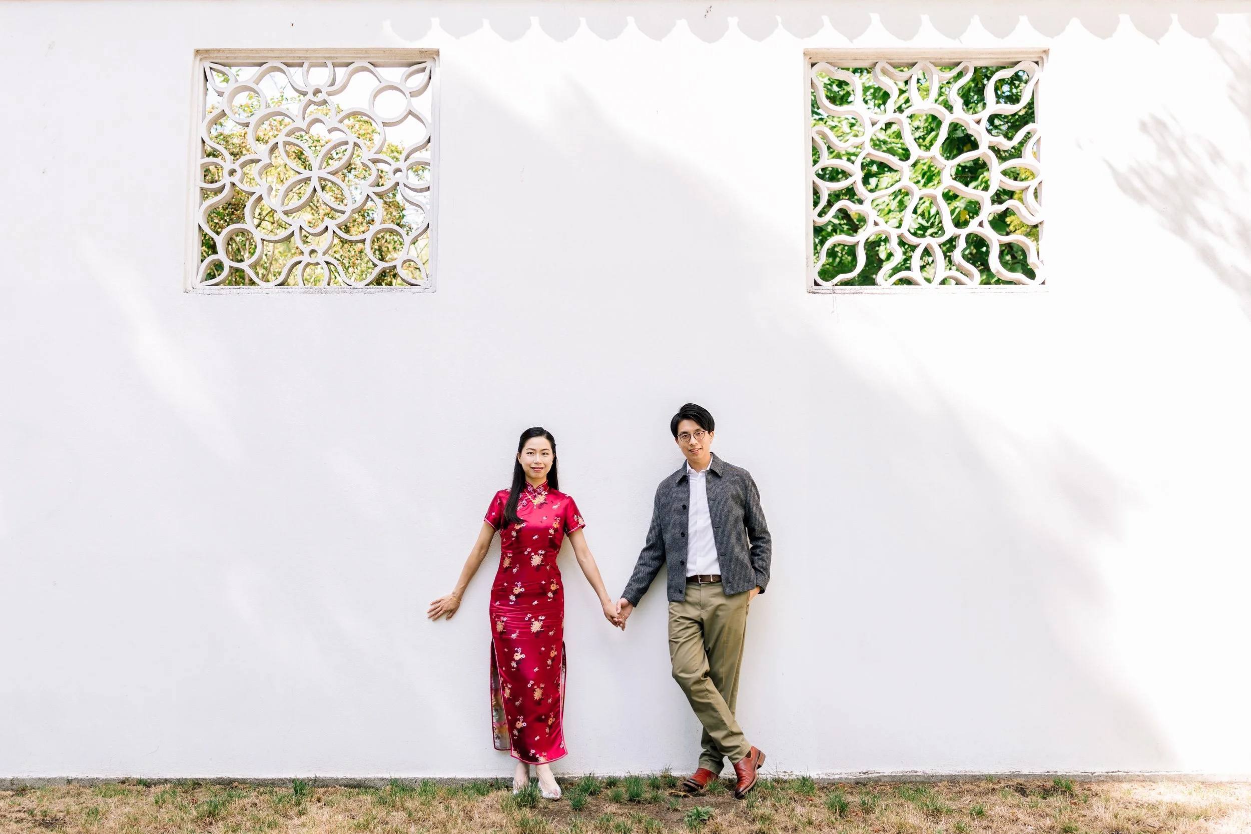 A man and woman holding hands, standing outdoors against a white wall with decorative Chinese lattice windows, the woman wearing a red Chinese dress and the man in a gray blazer and khaki pants.