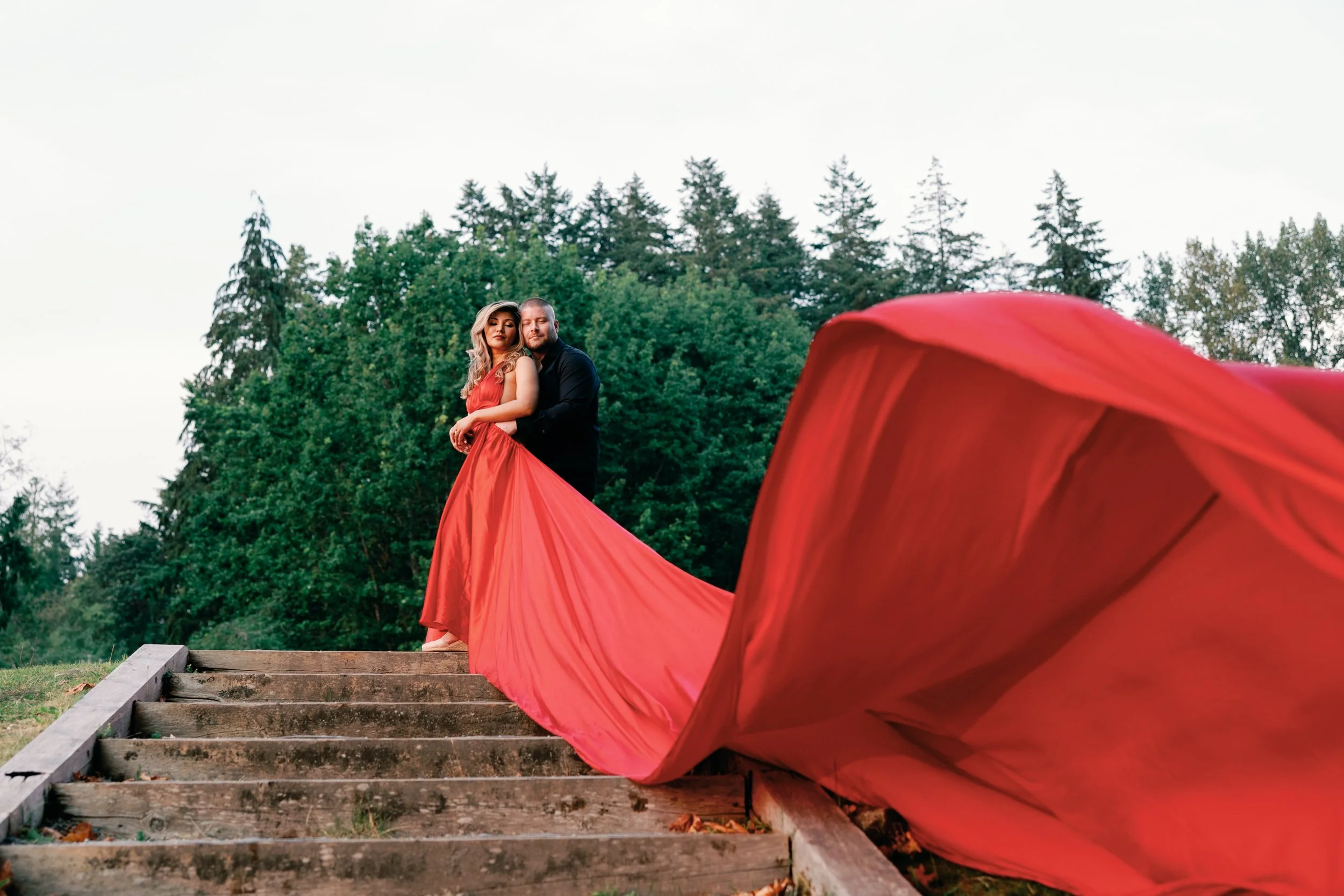 A couple in formal attire standing on outdoor wooden steps, with the woman wearing a long red gown and the man in a black suit, surrounded by green trees.