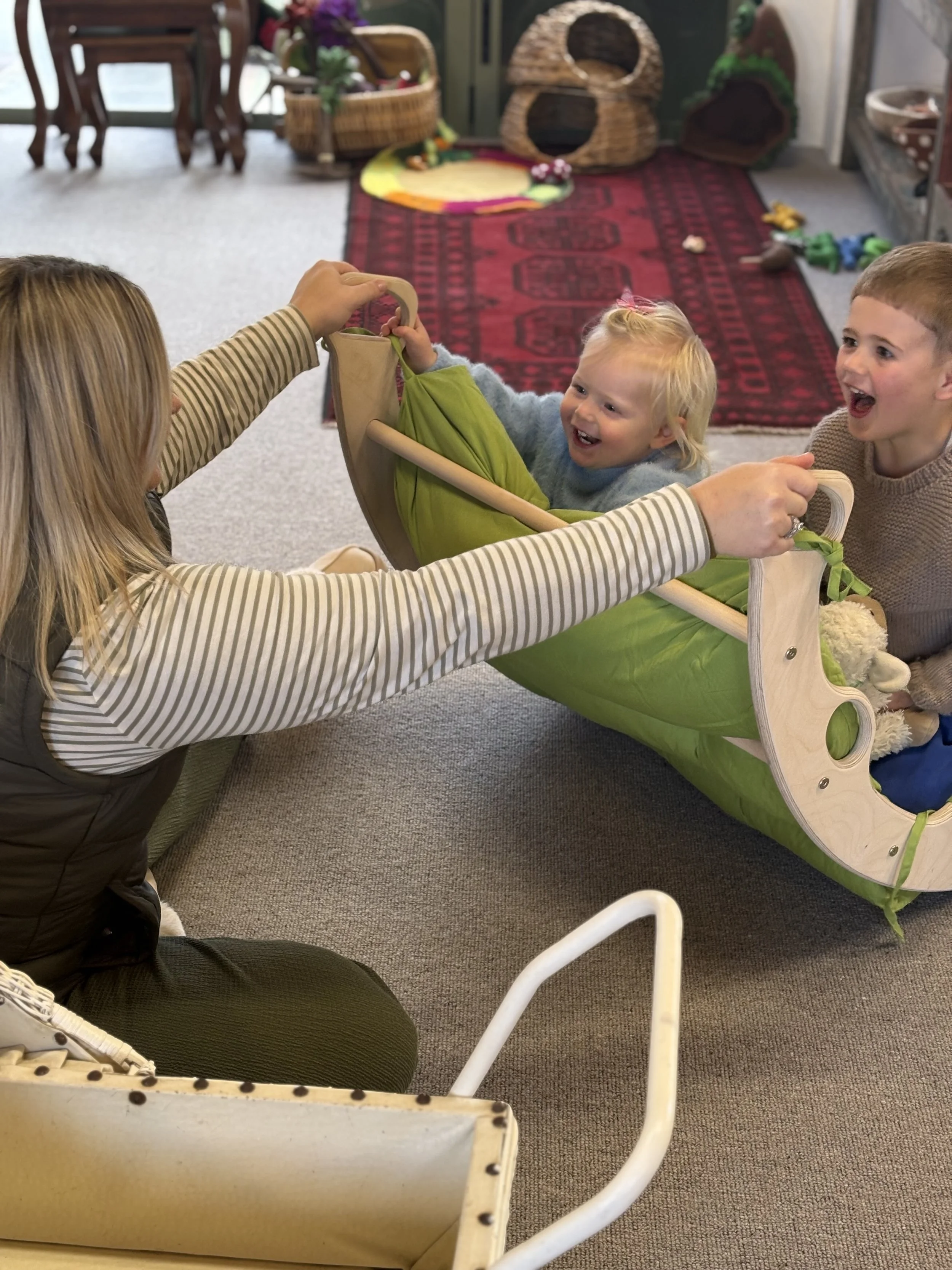 A woman and two children playing together with a wooden and fabric toy train on a carpeted floor in a room filled with pet toys and furniture.