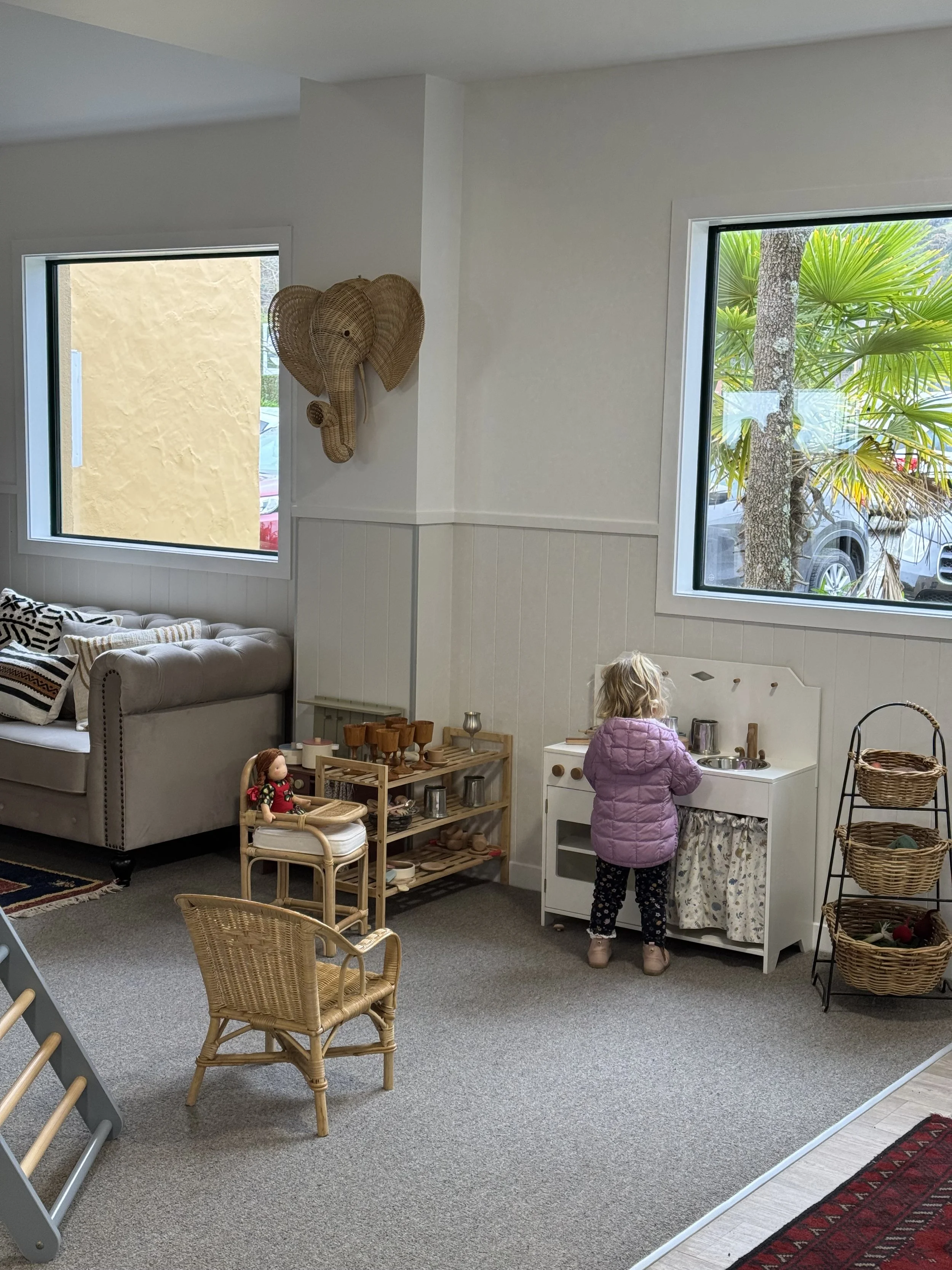 A young girl playing with a toy kitchen set in a cozy living room. There is a beige sofa with patterned pillows, a small wooden chair, and a doll on a stand nearby. Wicker baskets and a shelf with toy dishes and utensils are also visible, along with