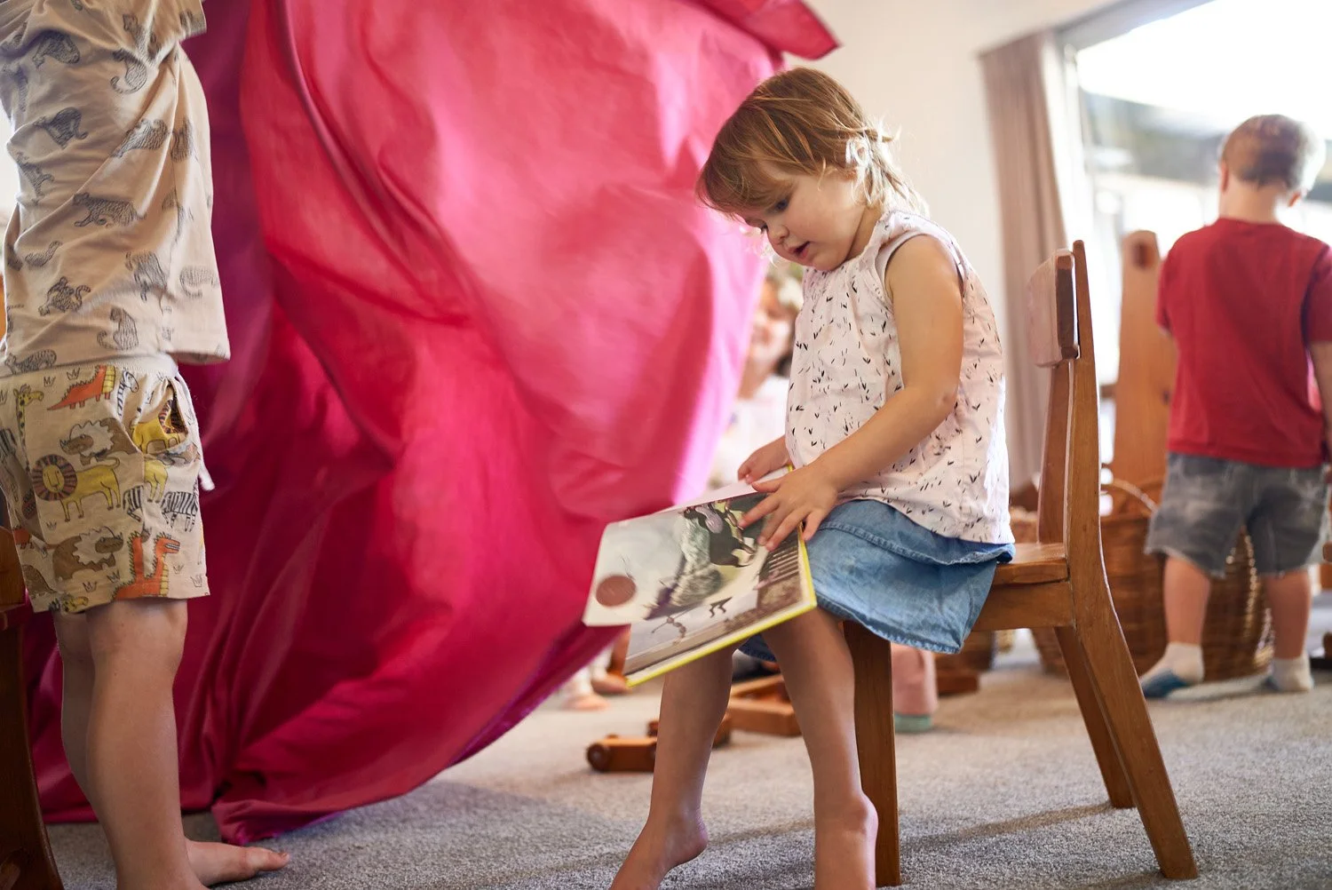 A young girl sitting on a wooden chair in a room, reading a book with pictures of animals, while other children are around.