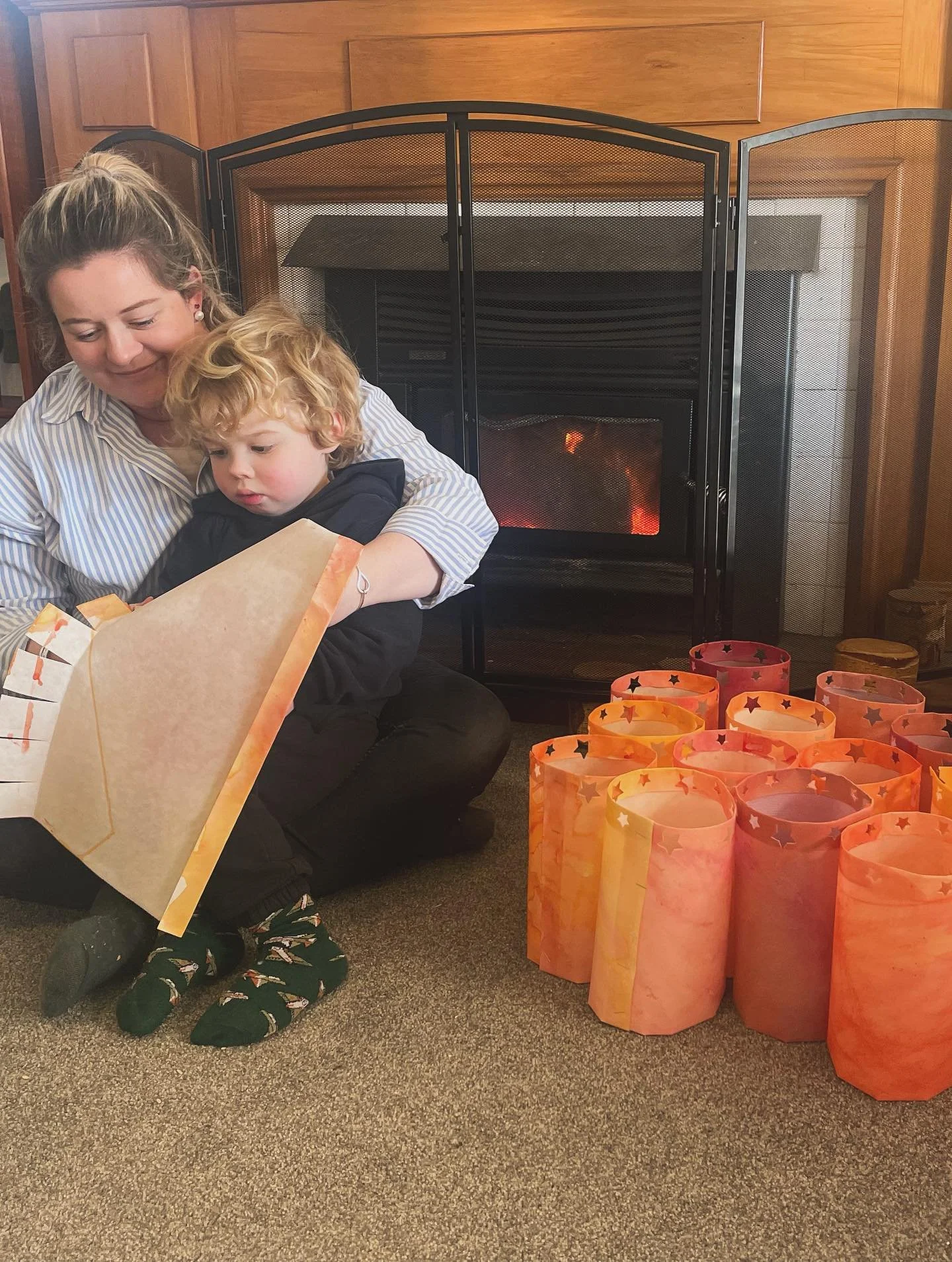 A woman and a young boy sitting on the carpet in front of a fireplace. The woman is helping the boy open a gift. There are orange paper lanterns with star cutouts on the floor beside them.