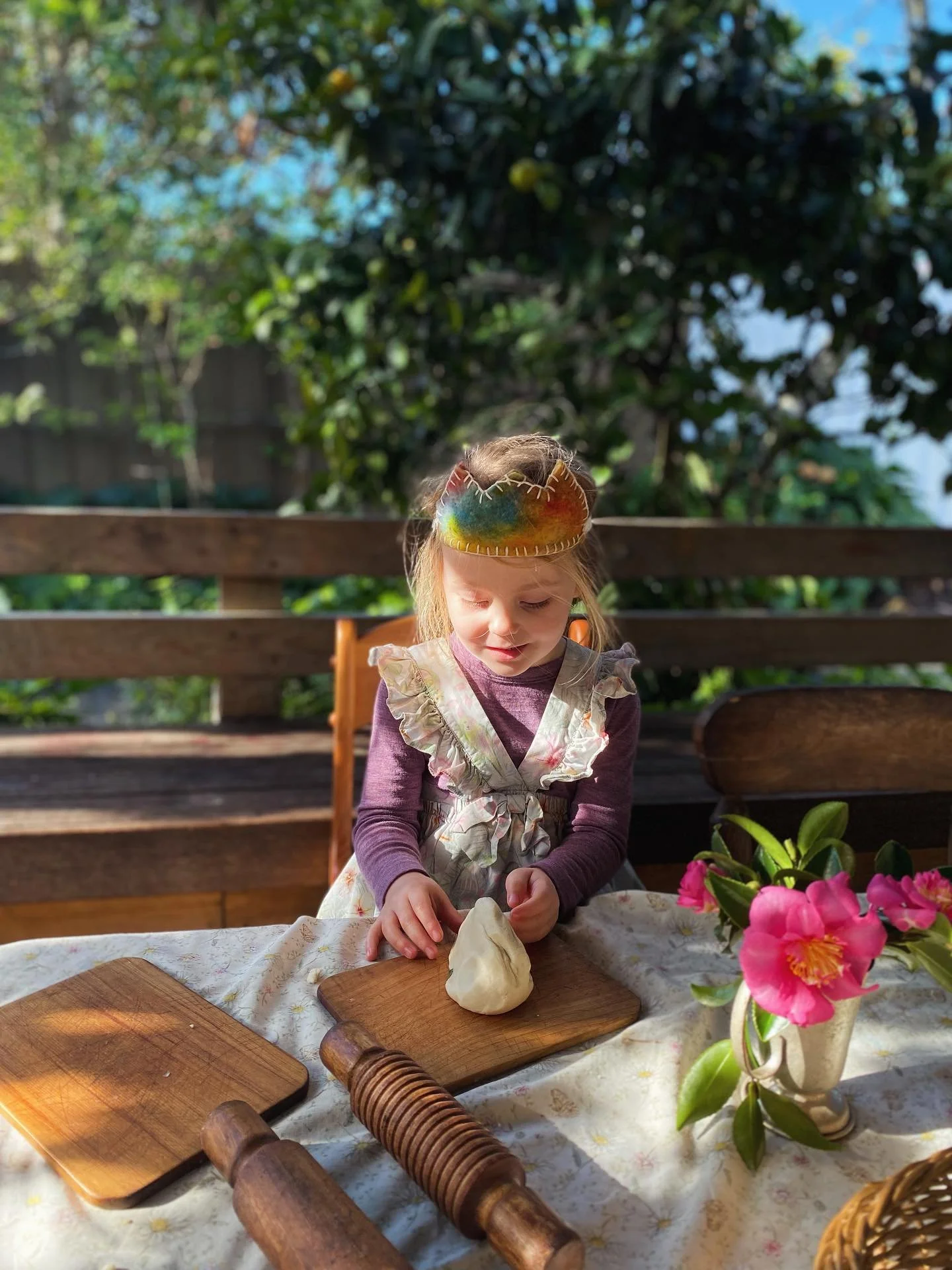 A young girl wearing a felted crown and ruffled apron, rolling dough on a wooden cutting board outdoors. There are pink flowers in a vase on the table, and a rolling pin nearby, with a wooden bench and green foliage in the background.