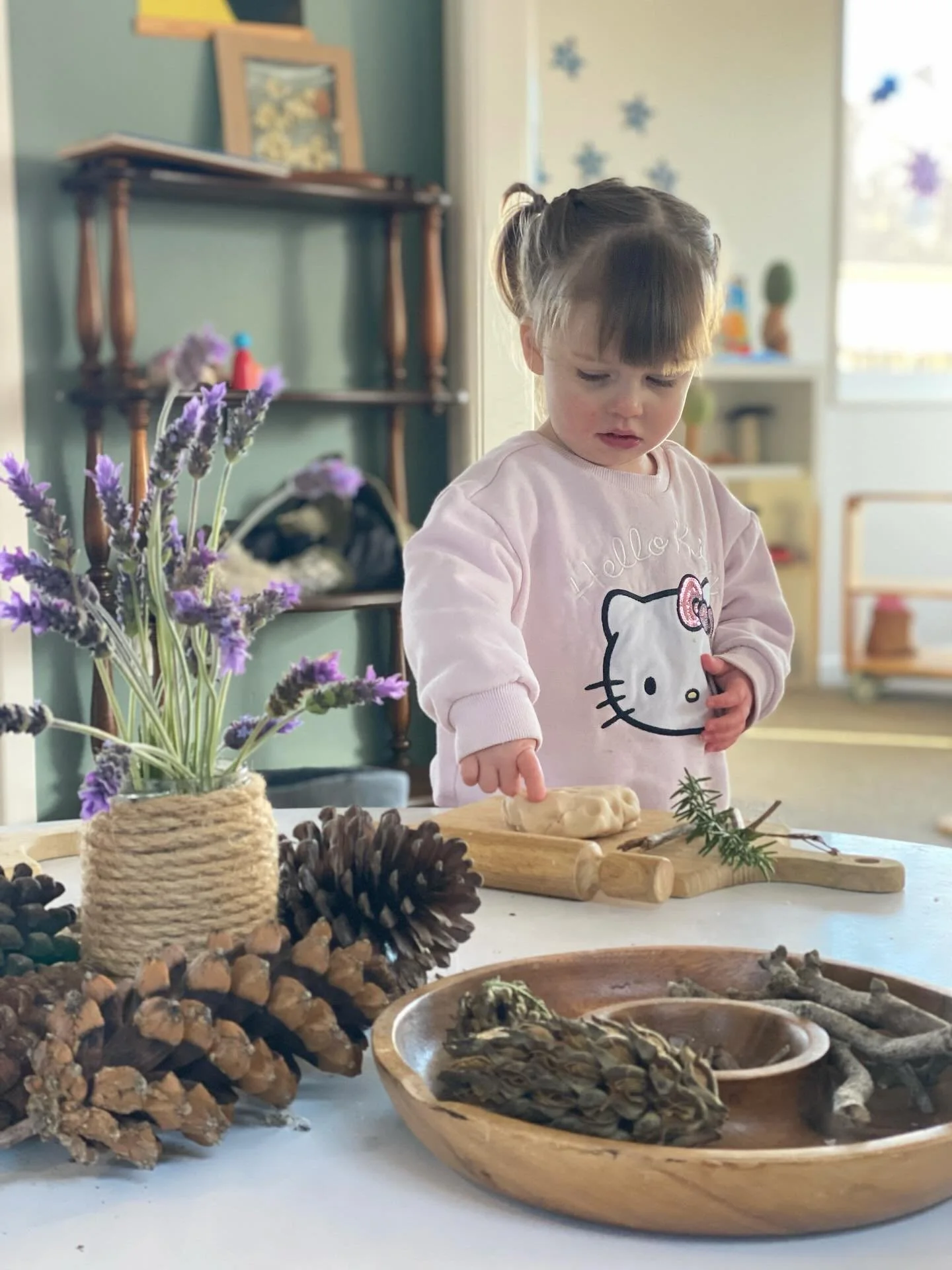 A young girl with brown hair in pigtails points to clay on a wooden cutting board. She wears a pink Hello Kitty sweatshirt. The table has pine cones, a wooden bowl with pine cones and twigs, a vase with purple flowers, and other natural objects. The