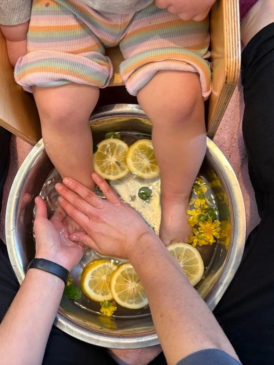 A child's legs sitting in a metal foot spa filled with water, lemon slices, and yellow flowers, with a parent's hands holding the child's feet.