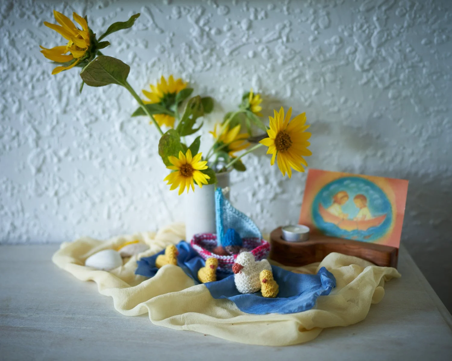 A small still life arrangement featuring a white vase with yellow flowers, a crocheted white dove, candles, a colorful drawing of two children in a boat, and decorative fabric on a table with a textured white wall background.