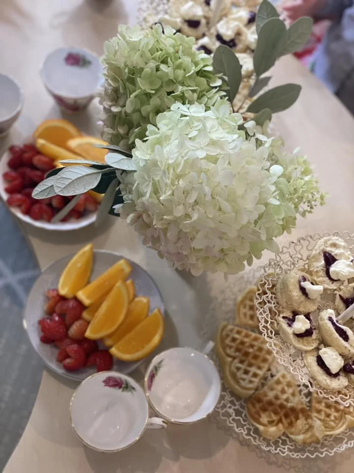 Close-up of a floral centerpiece with white and light green hydrangeas, surrounded by fruit plates with orange slices, strawberries, and raspberries, along with waffle cones and small cups.
