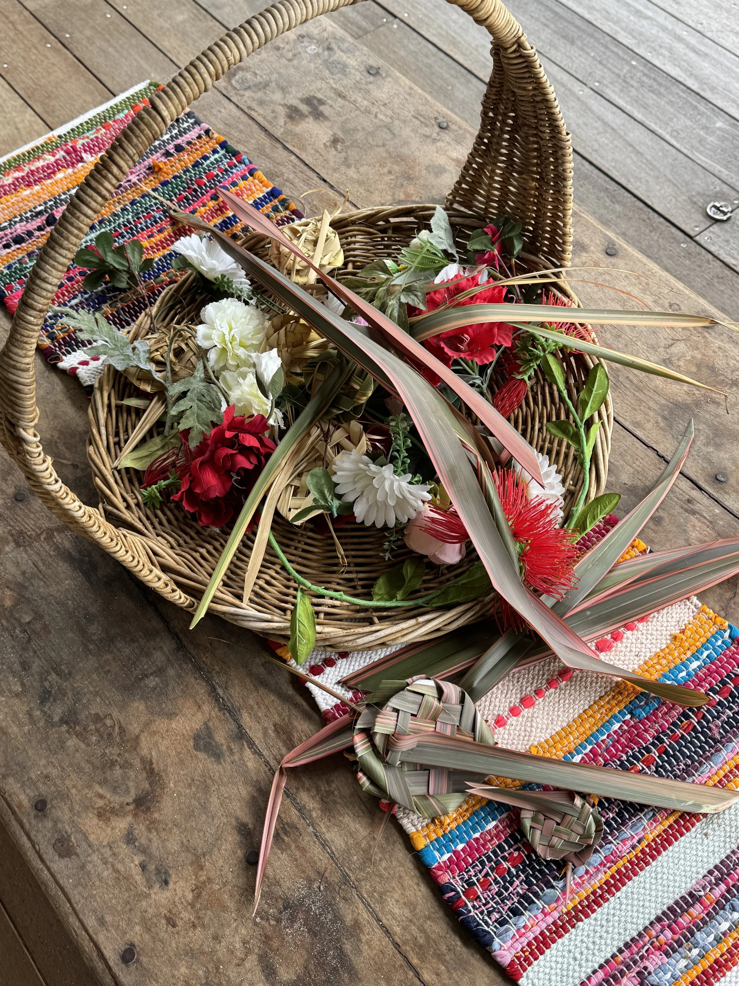 A wicker basket filled with assorted flowers and leaves, placed on a multicolored woven mat on a wooden surface.