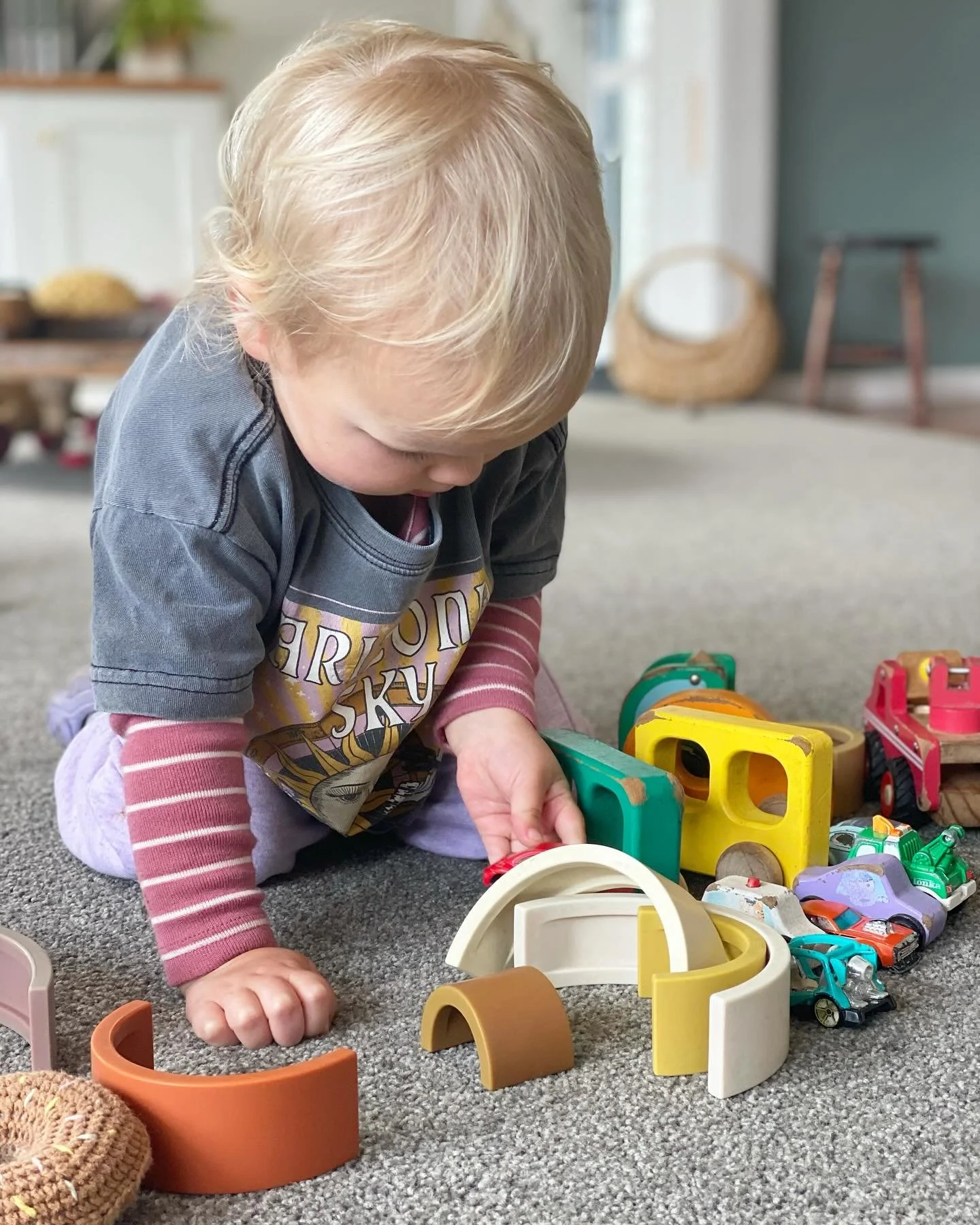 A young child playing with colorful toy blocks and cars on a carpeted floor in a room with a basket and furniture in the background.