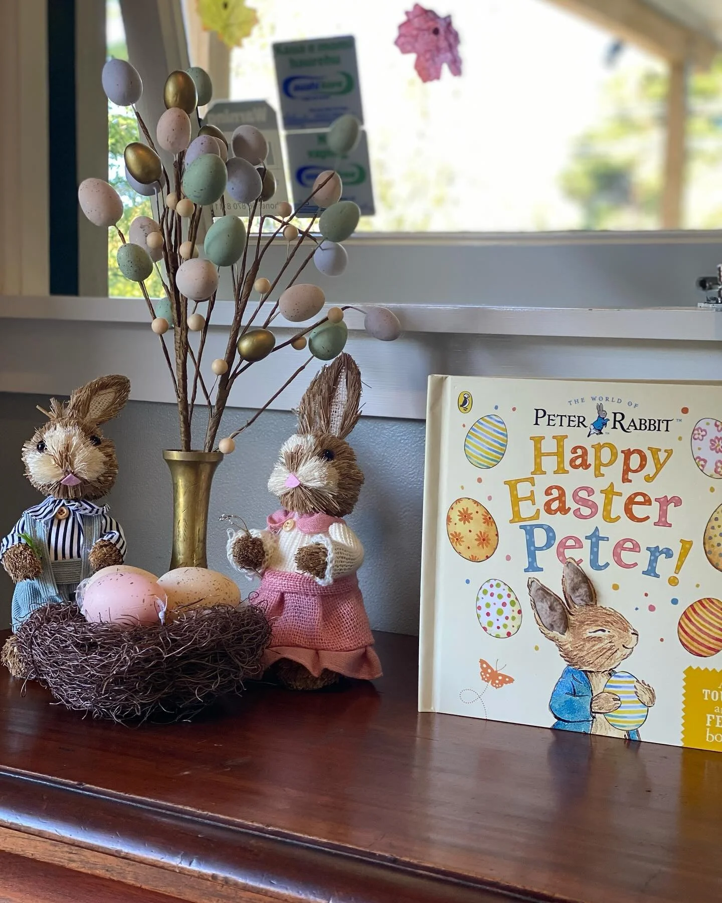 Decorative Easter display with plush bunny dolls, a branch with pastel-colored eggs in a gold vase, an Easter-themed book titled 'Happy Easter Peter!' by Peter Rabbit, and a window with green foliage outside.