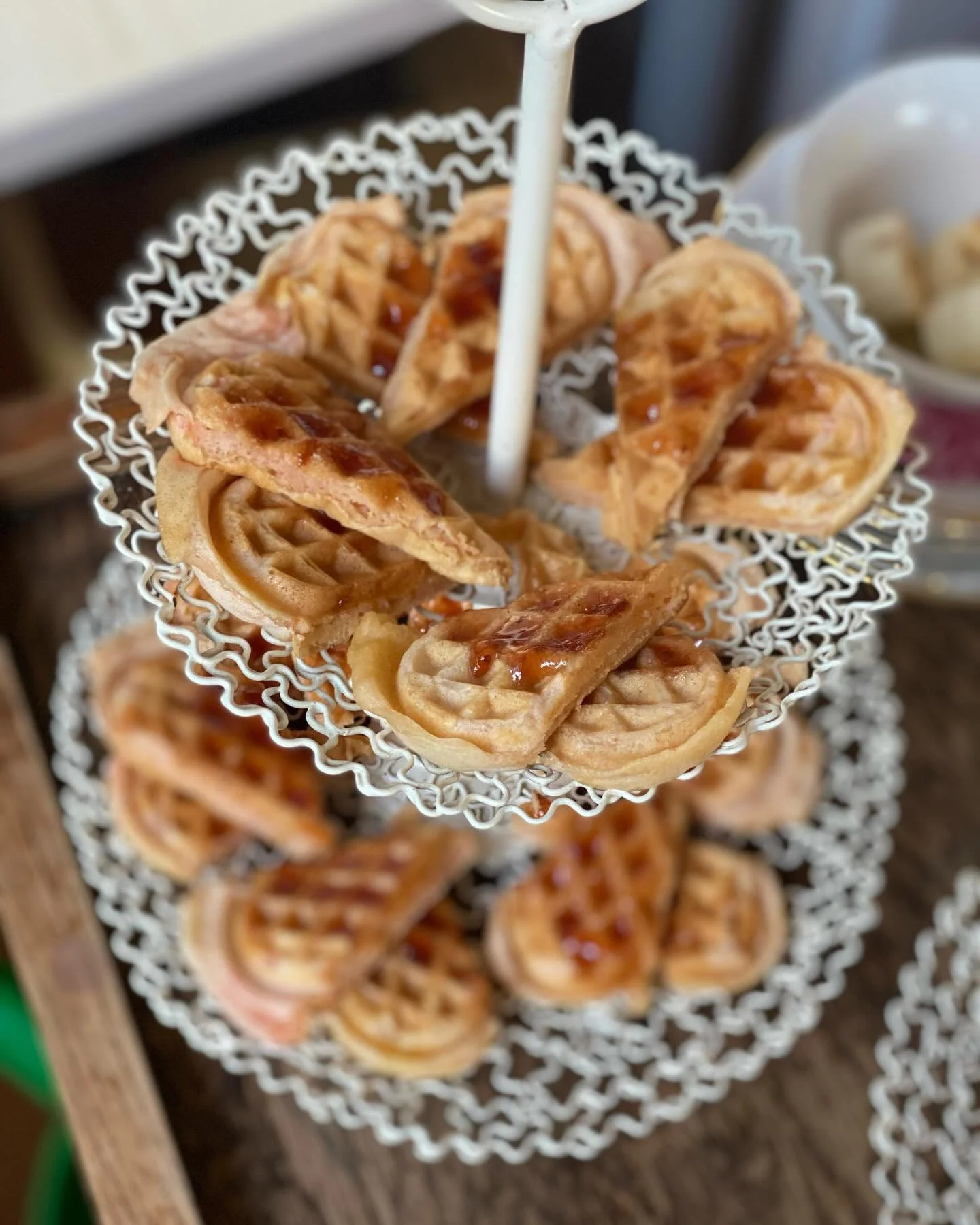 Two-tiered metal serving stand with mini waffles topped with syrup, on a rustic wooden table.