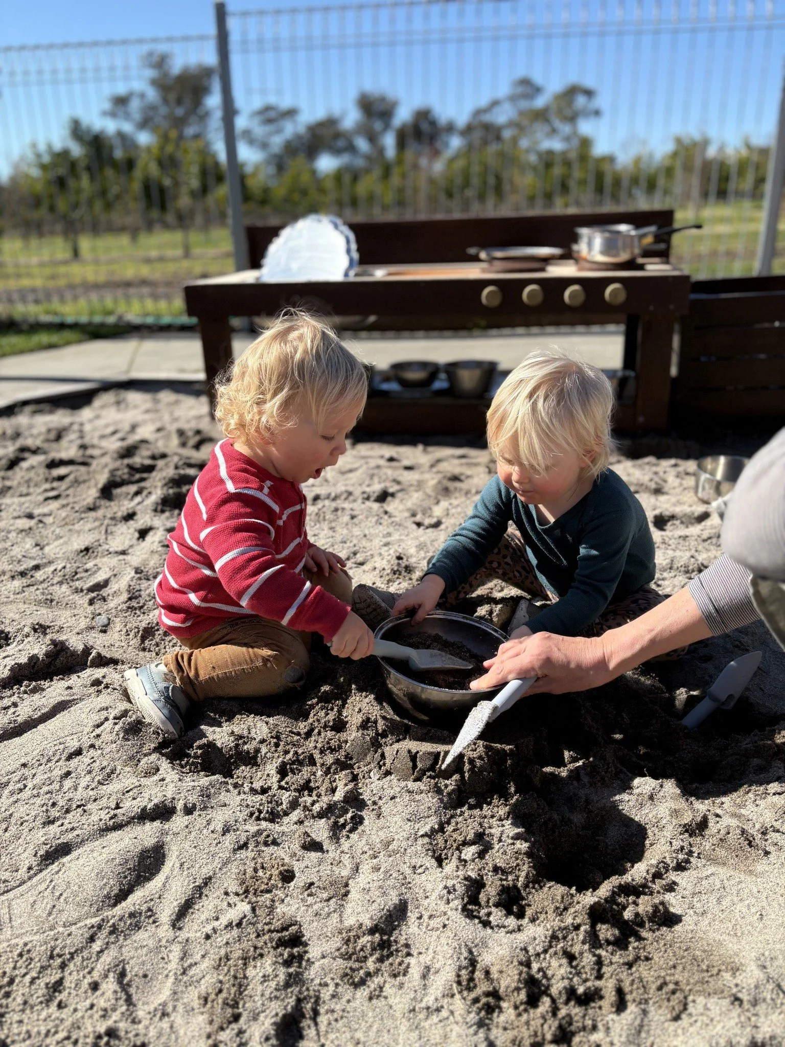 Two young children playing in a sandbox with a woman, using shovels and a bowl, outdoors on a sunny day with a fence and garden in the background.