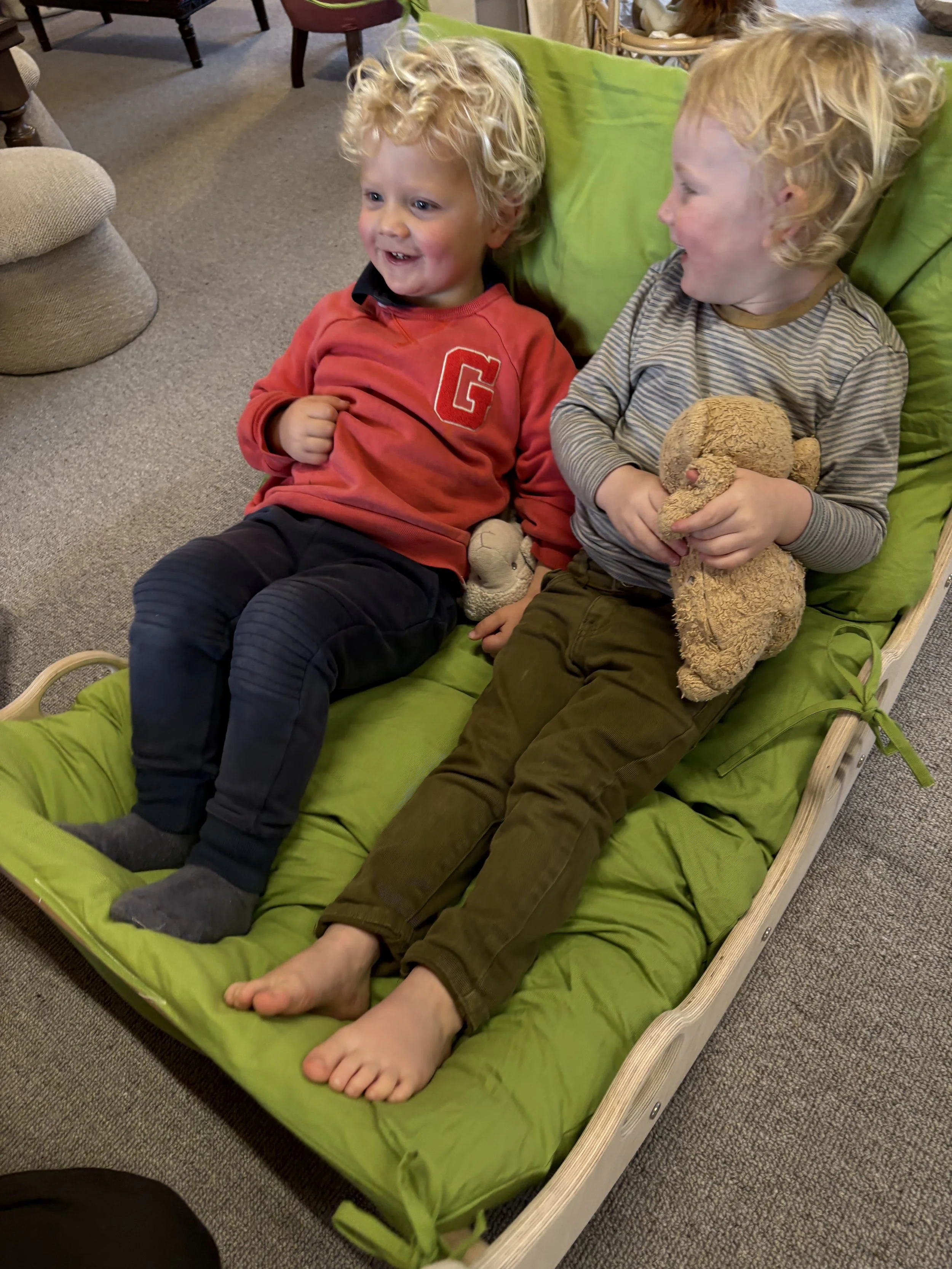 Two young children with curly blonde hair sitting on a green cushioned lounge, smiling and laughing, one holding a teddy bear.