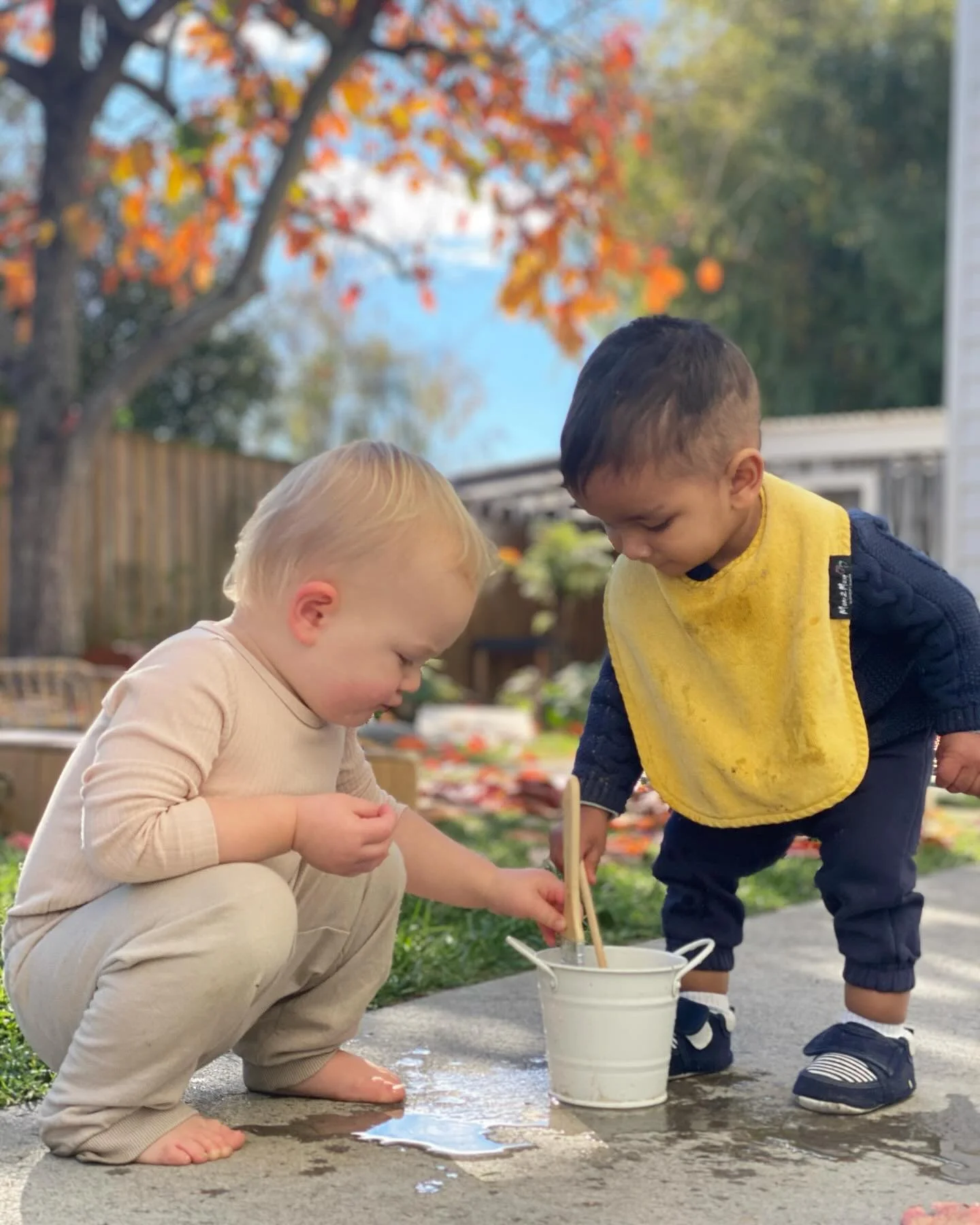 Two young children playing outside on a concrete path with a white bucket, with autumn-colored trees and a clear blue sky in the background.