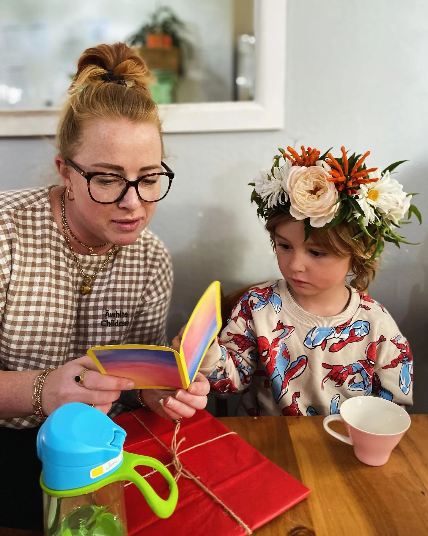 A woman with glasses and a young girl wearing a flower crown sit at a table, looking at a colorful card together. The woman has her hair in a bun, and the girl is wearing a Spiderman shirt. There is a pink cup and a blue water bottle on the table, al