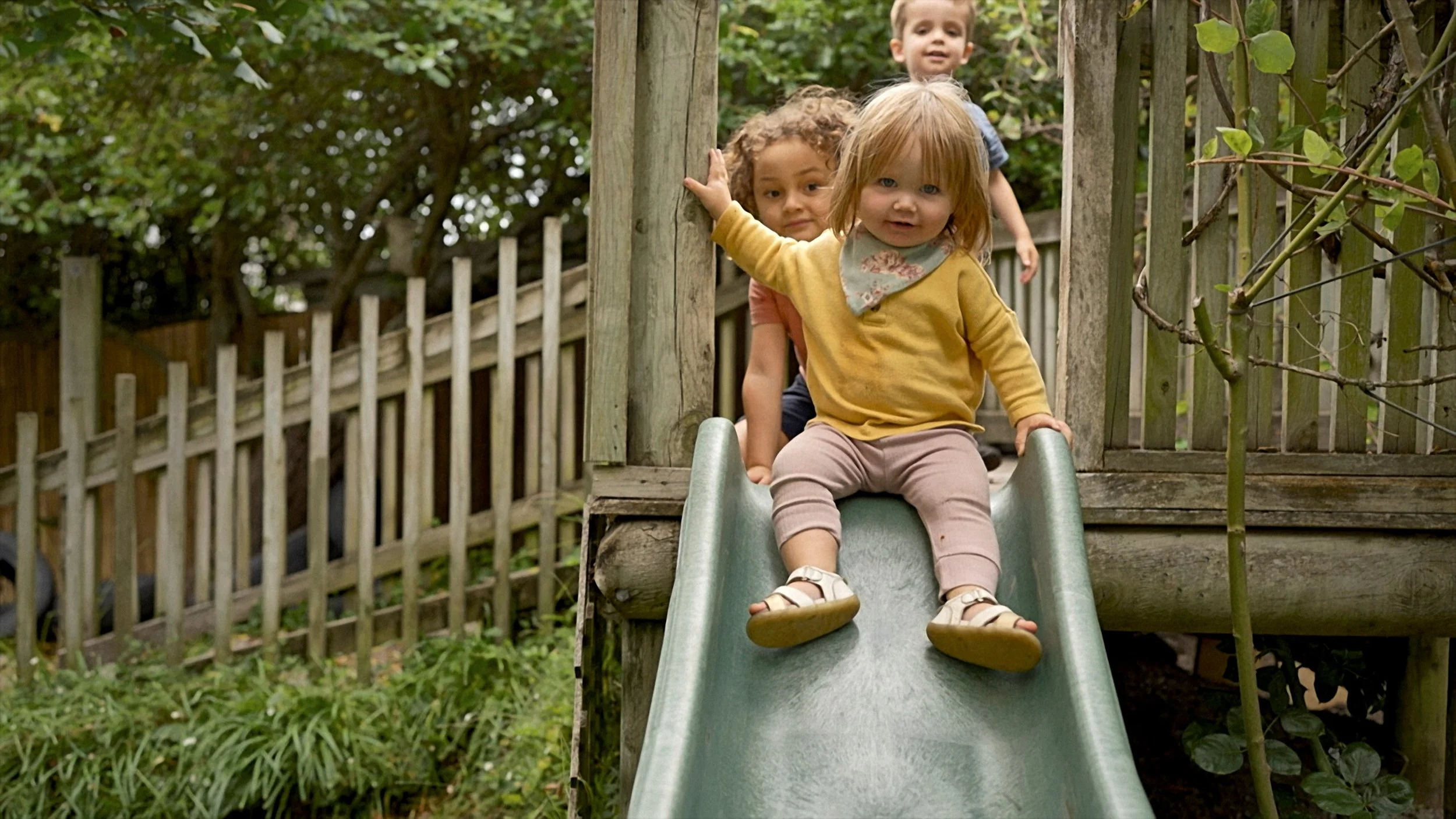 Kids playing on a slide in a backyard with a wooden fence and trees in the background.