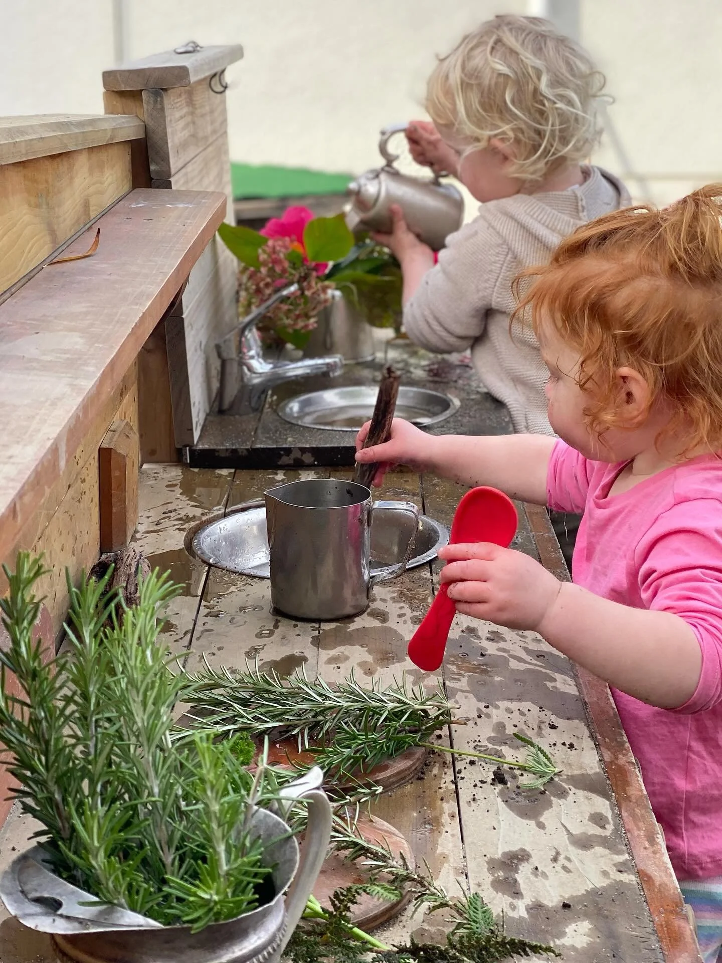 Two children with curly hair are playing at an outdoor pretend kitchen. The girl in the pink shirt holds a red spoon and is stirring a mug, while the boy with blonde hair in a beige sweater is pouring water from a metallic kettle. The pretend kitchen