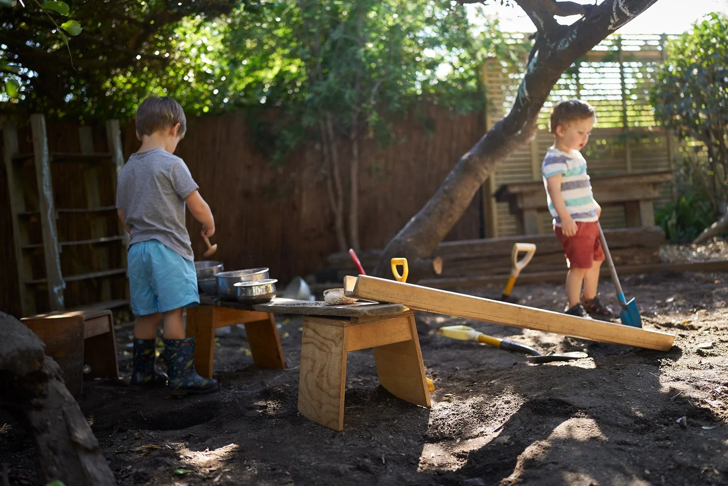 Two young boys are playing outdoors in a backyard, engaging in pretend construction work. One boy is near a small wooden table with metal pots, wearing a grey t-shirt, blue shorts, and rubber boots. The other boy, in a striped t-shirt and red shorts,