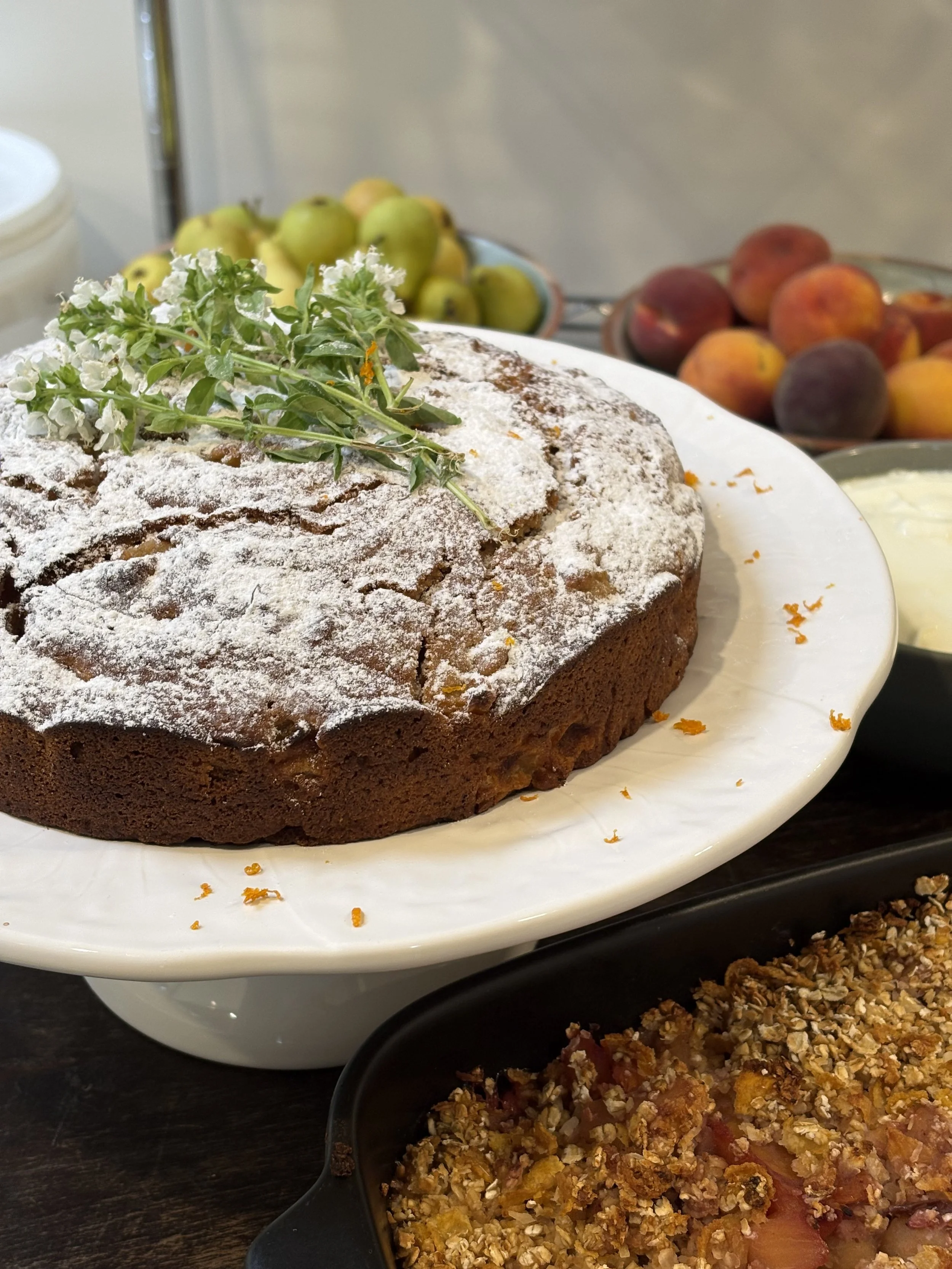 Chocolate cake dusted with powdered sugar, garnished with a sprig of herbs, with bowls of green apples, peaches, and baked fruit crisp in the background.