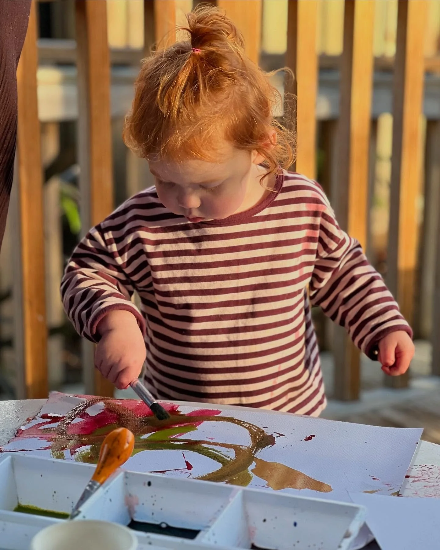 A young girl with red hair, wearing a striped long-sleeve shirt, painting with watercolors on a piece of paper outside on a wooden deck.