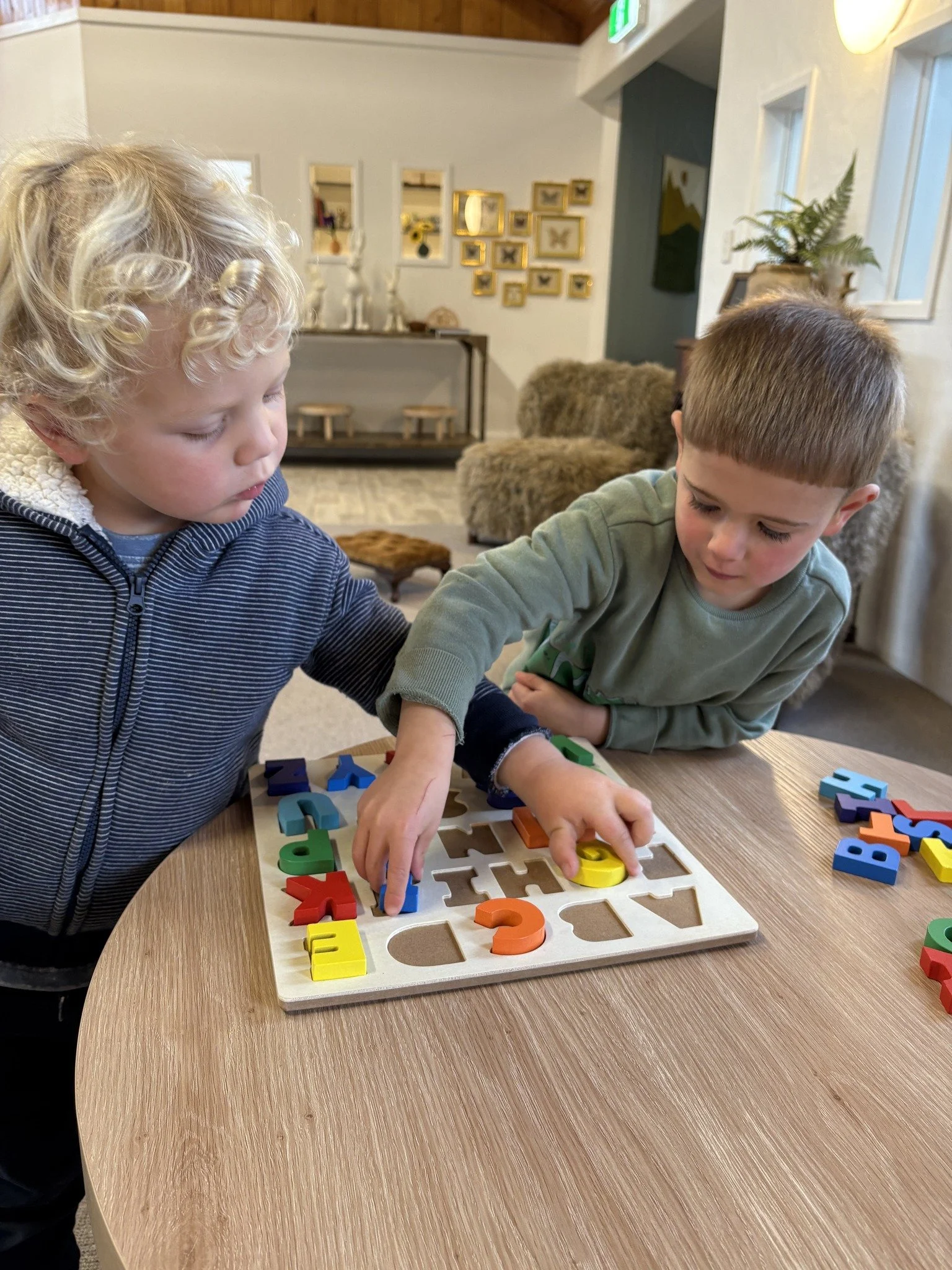 Two young boys playing a puzzle game with colorful number and letter pieces on a wooden table in a cozy living room.