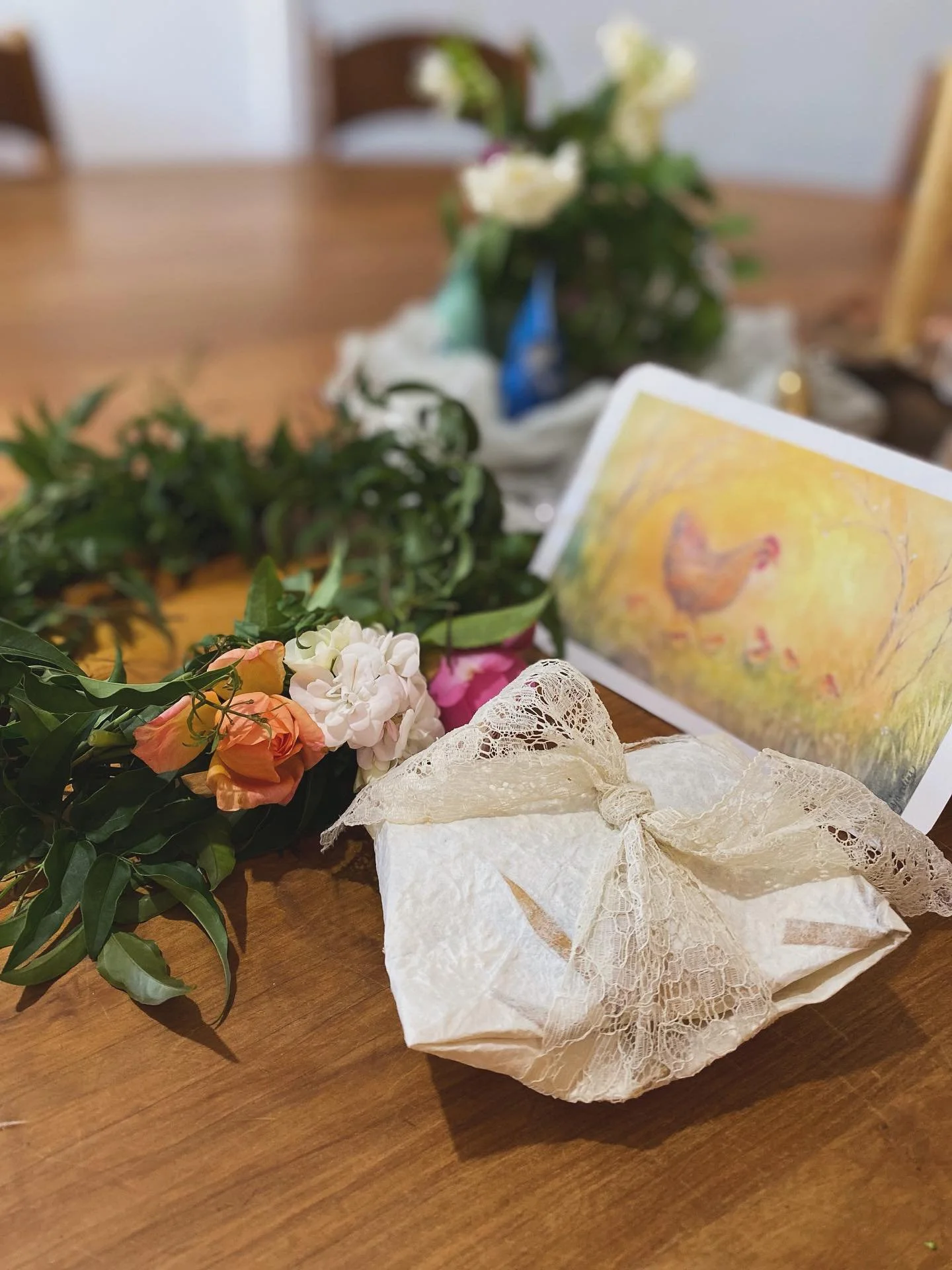 Decorative table setting with a wrapped gift, fresh flower crown, and a colorful chicken-themed card on a wooden table.
