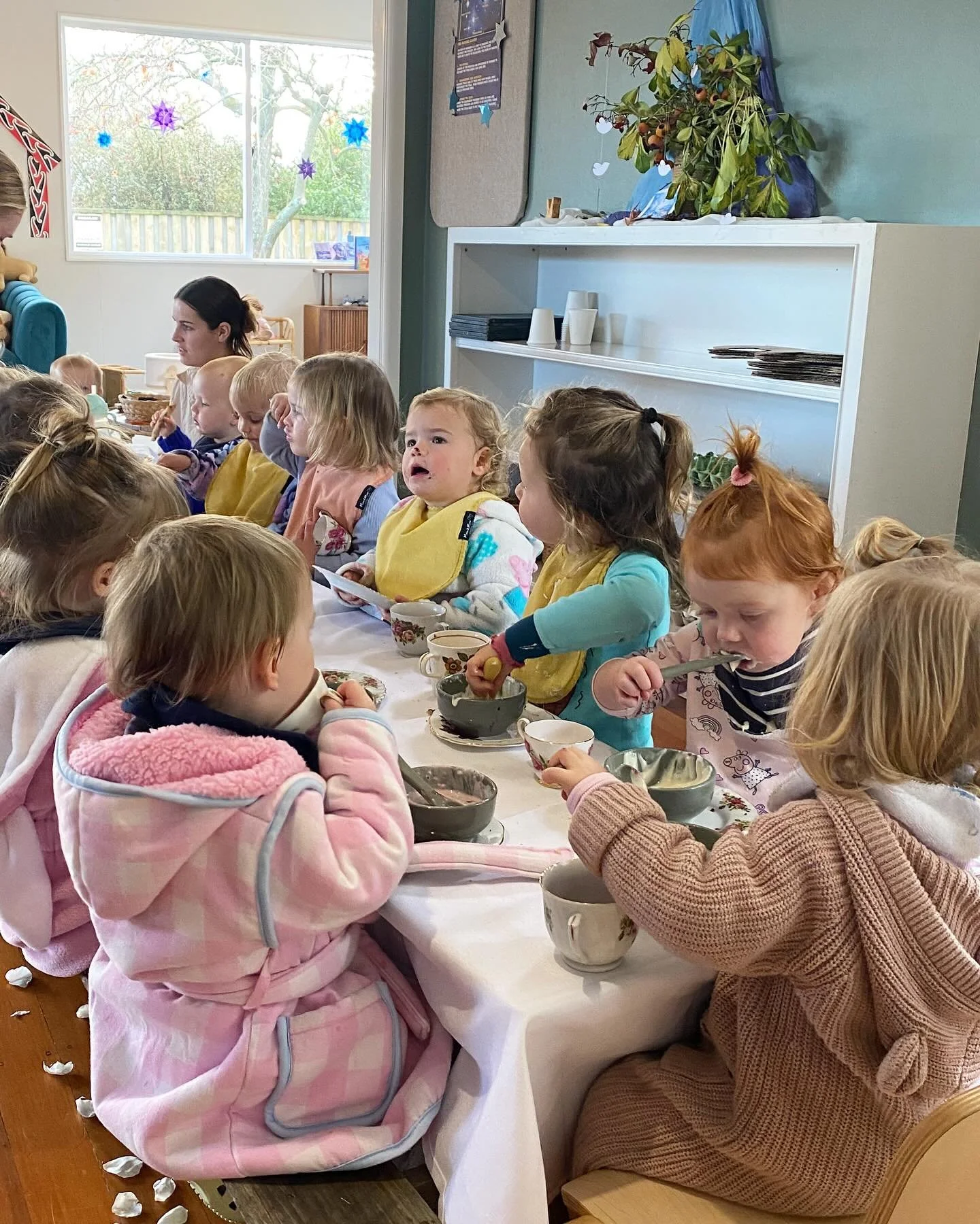 Children sitting at a long table having snack or meal, some with bowls and cups, in a room with a large window, decorated with paper snowflakes and a plant on a white shelf.