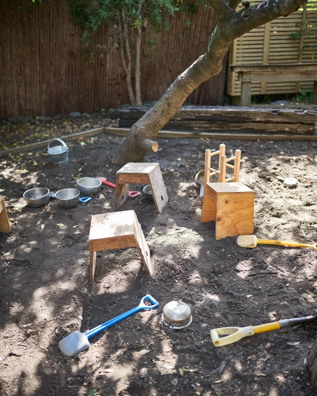 Children's outdoor play area with small wooden stools, colorful shovels, metal bowls, and a sandcastle mold, surrounded by dirt, trees, and a fence.