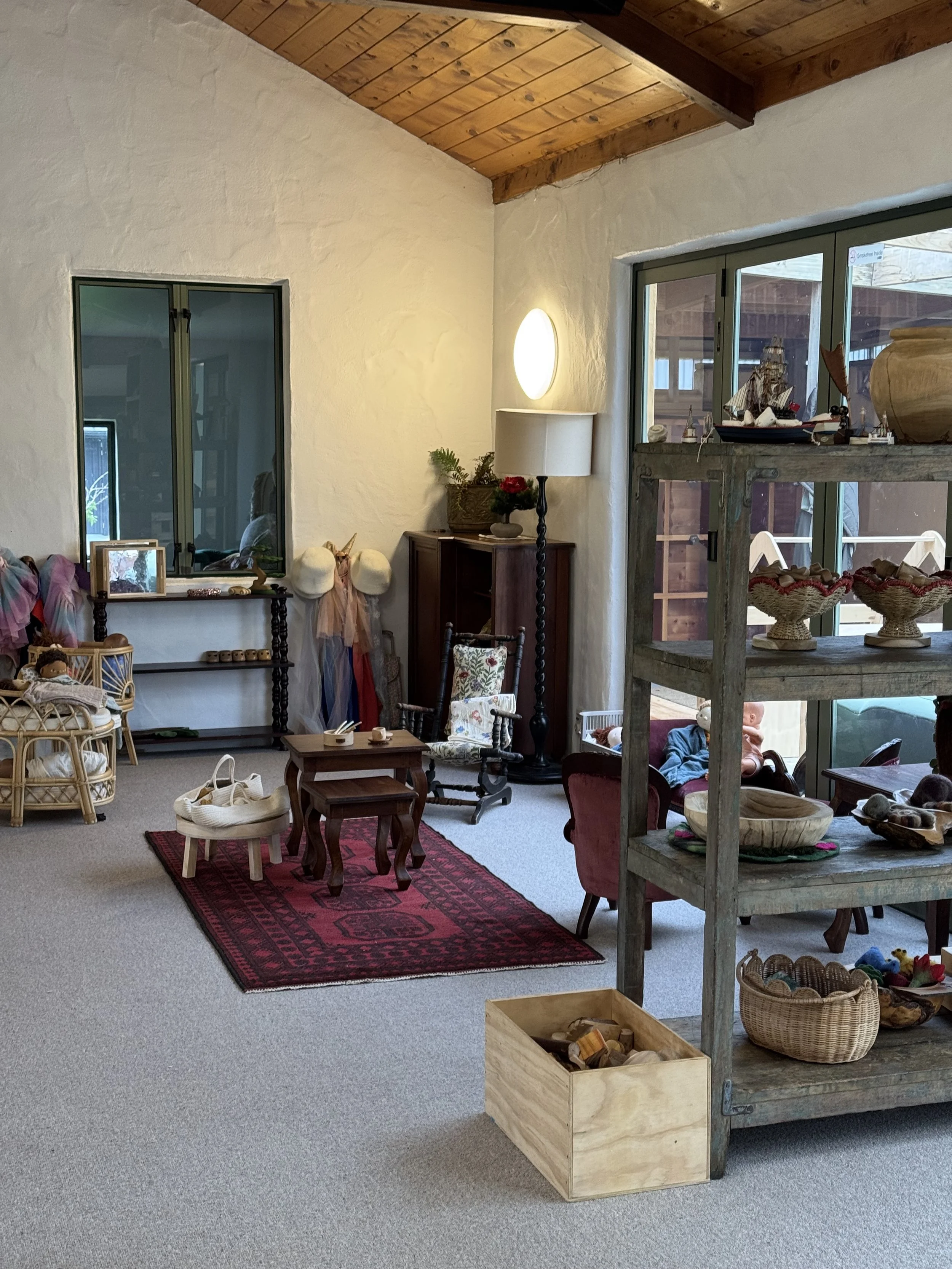Living room with high wooden ceiling, white textured walls, and large windows. Contains a wooden rug, small tables, chairs, shelves with decorative items, and toys.