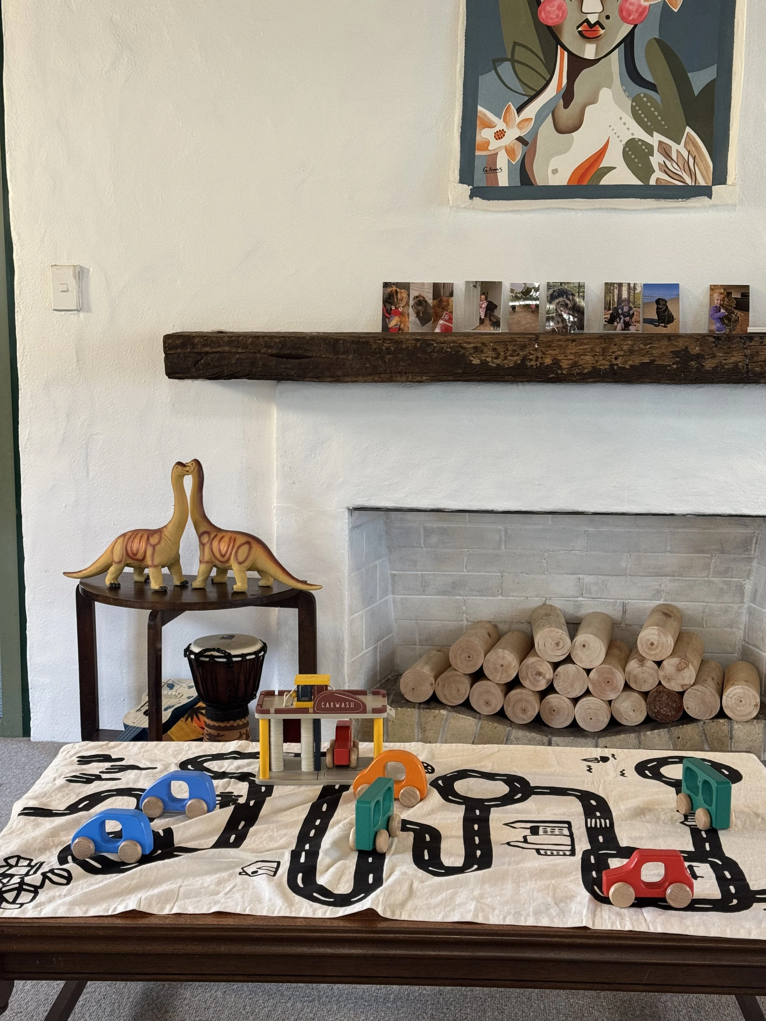 A children's play area with a play mat featuring black road illustrations, small colorful toy cars, a toy gas station, and a toy traffic cone. Behind the play area, there is a fireplace with a stack of wooden logs and a wooden beam mantel. To the lef