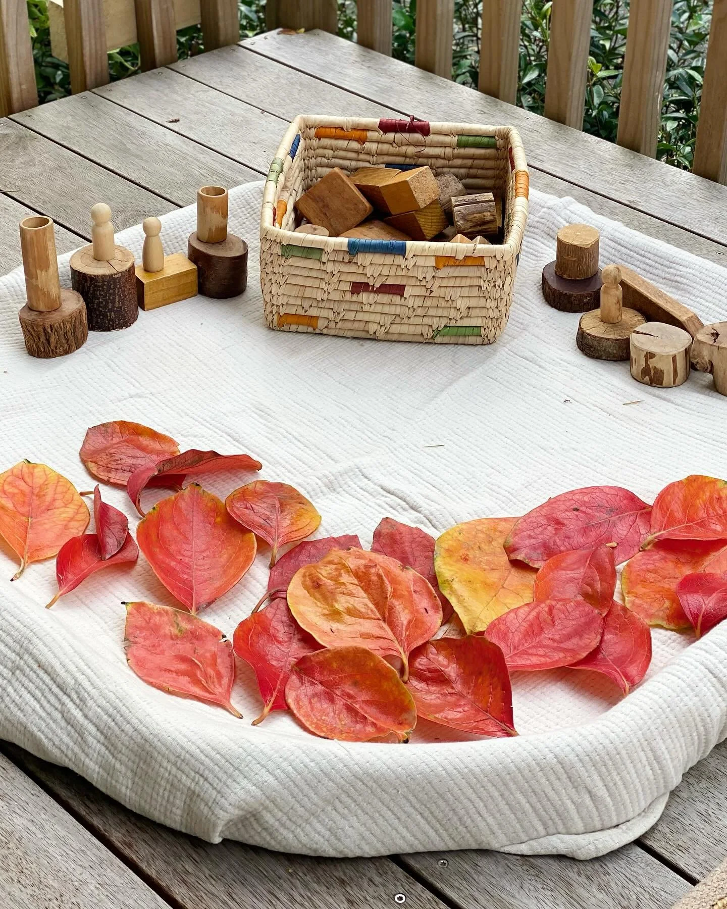 A table with a white cloth, a basket of wooden blocks, and a circle of red and orange leaves.