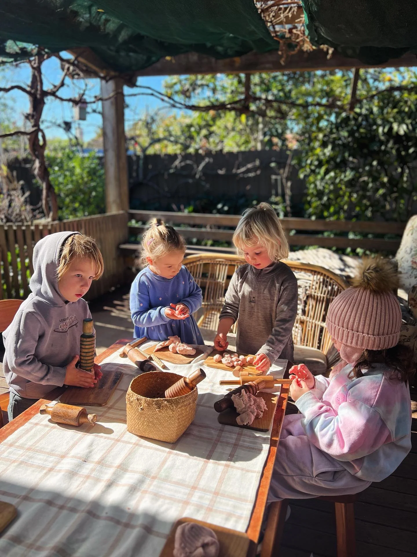 Four children cooking or preparing food outdoors at a wooden table under a canopy, with trees and a fence in the background.