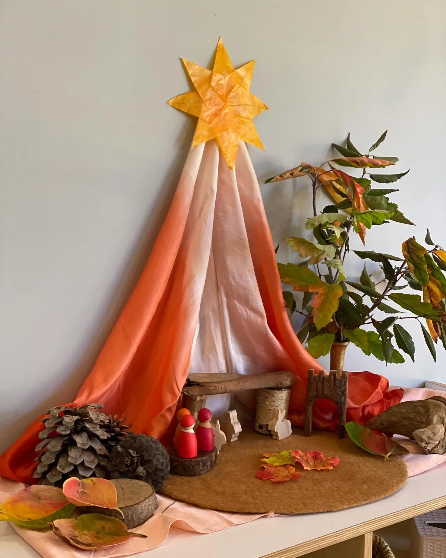 Decorative autumn scene with orange fabric draped to resemble a tent, topped with a yellow paper star. Includes pinecones, small toy figurines, wooden logs, a potted plant, and scattered fall leaves on a brown circular mat.