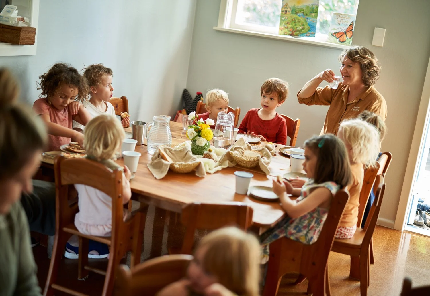 A group of children and an adult woman gathered around a wooden table for a meal in a cozy room with a window. The woman appears to be giving a toast or drink to the children, who are sitting on wooden chairs. The table is set with plates, cups, a ce