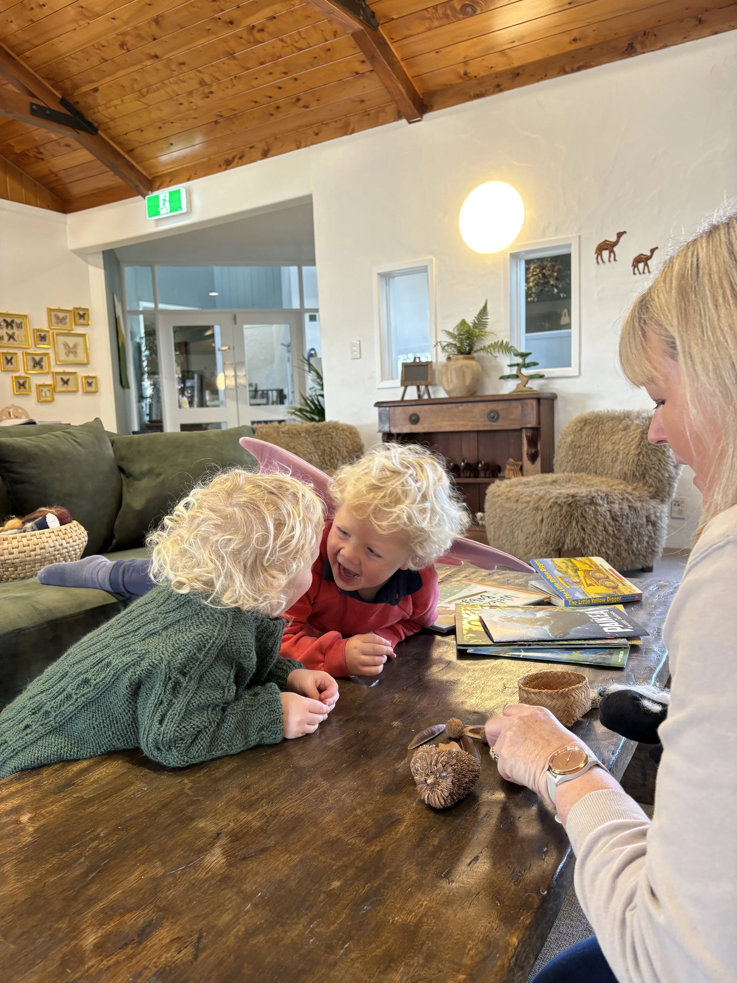 Two children with blonde curly hair and a woman with blonde hair sitting at a wooden table, engaging in a playful and happy moment. The children are leaning towards the woman, smiling and talking. The woman has a watch on her wrist and is holding som
