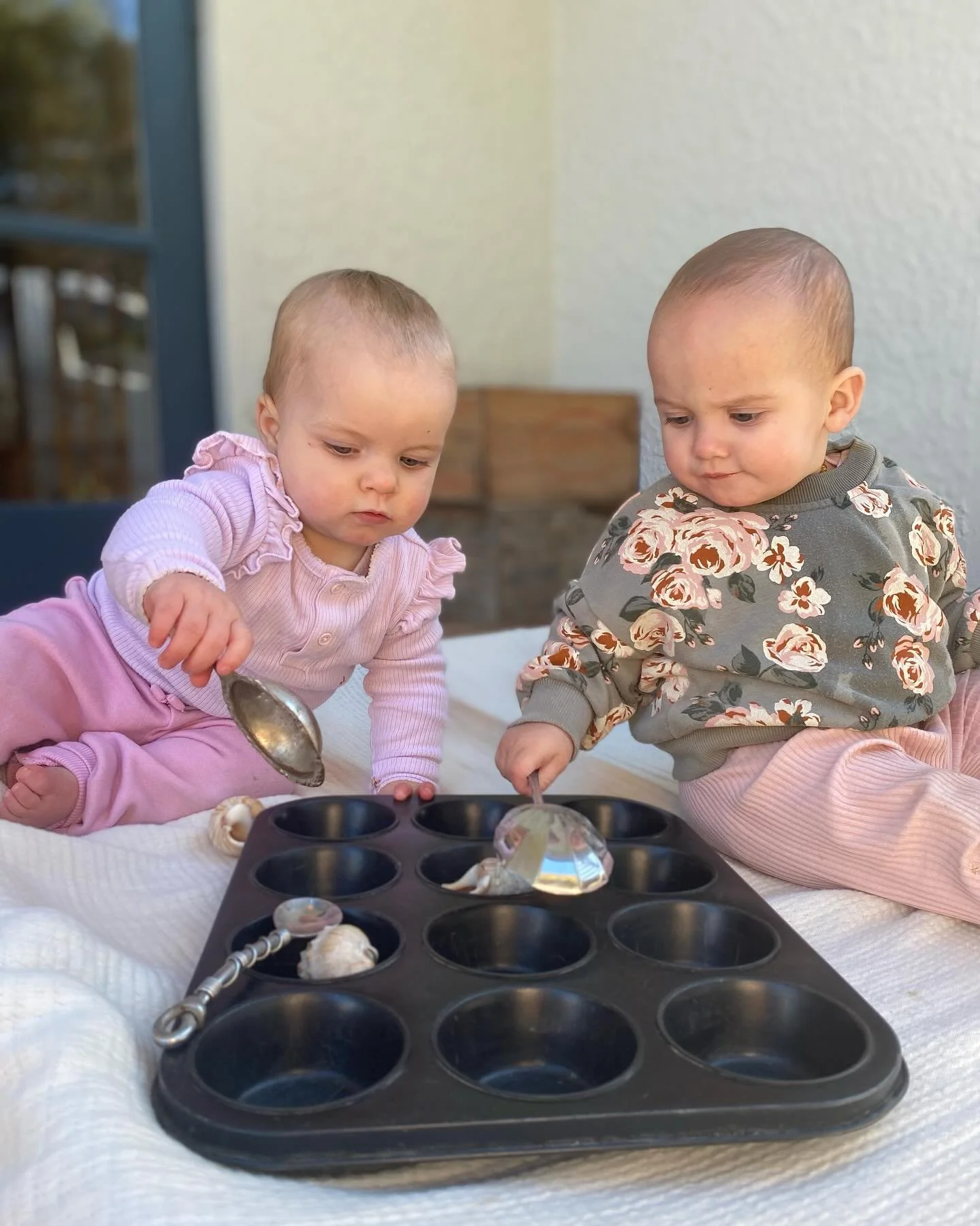 Two young children playing with a muffin tin and shiny toys on a bed or soft surface outside.