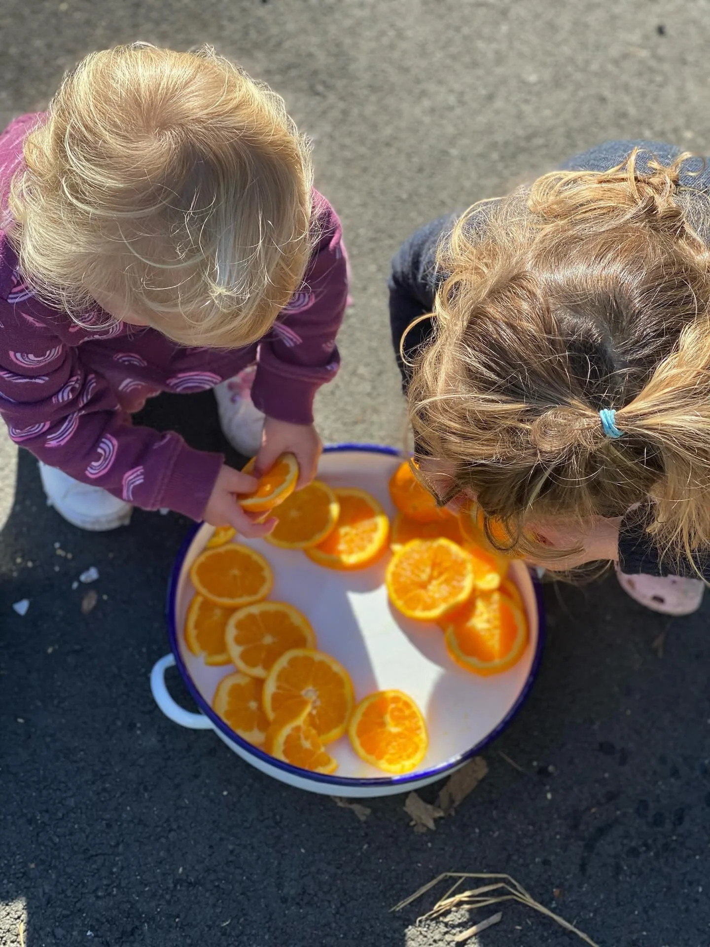 Two young children with blonde hair bending over a large bowl of sliced oranges on a paved surface.