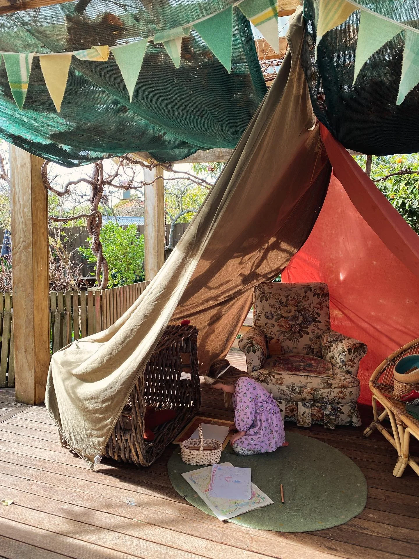 A young girl with long hair, dressed in pink pajamas with floral patterns, kneeling on a small green rug outdoors, drawing with colored pencils on paper, under a makeshift tent made of fabric and bunting flags, with outdoor furniture and trees in the