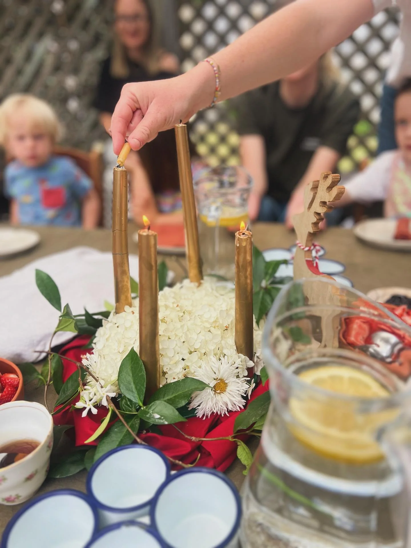 Three young children inside a house near a doorway leading to an outdoor patio, with an adult woman outside. The children are engaging in play or activities near a wooden table, and the scene is bright with natural sunlight.