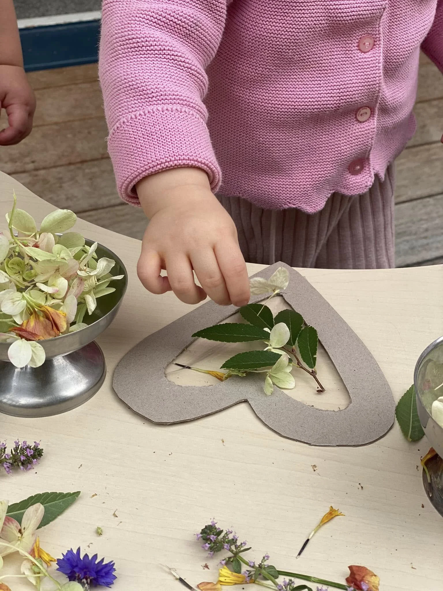 A child in pink sweater and striped pants arranging flowers on a wooden table for a craft project.