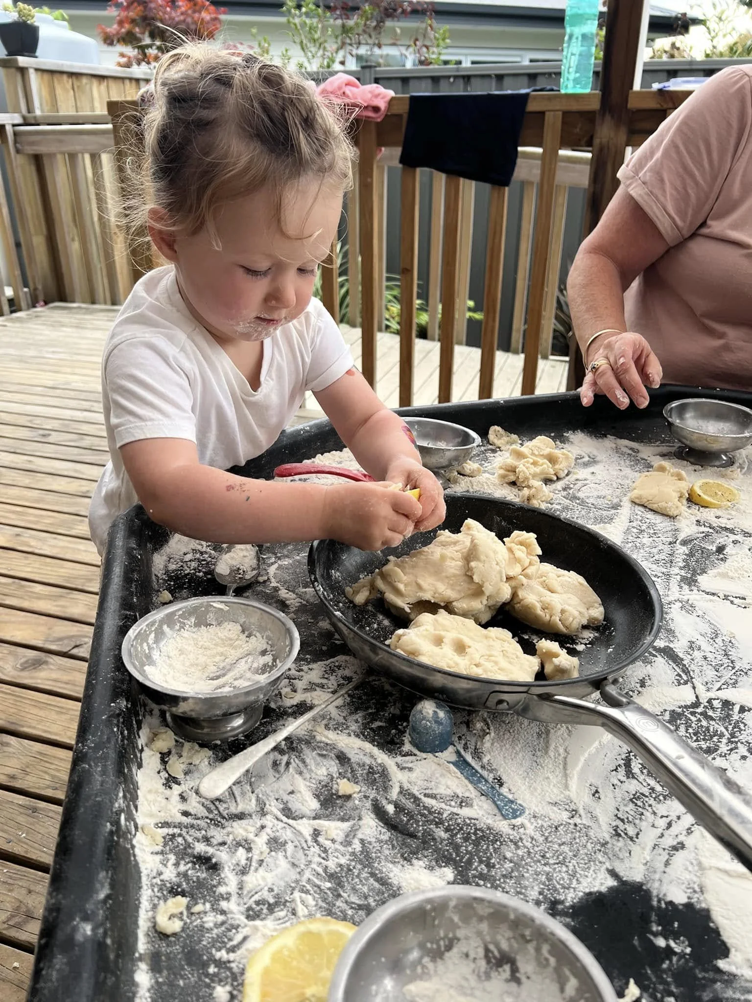 A young girl with curly hair and a white shirt is playing with dough at an outdoor table on a wooden deck. There are bowls, lemon slices, and other baking tools on the table, and an adult is assisting nearby.