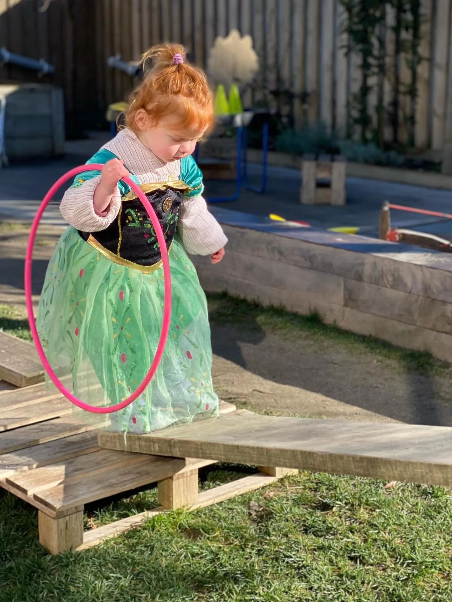 A young girl dressed as a princess with a green and black dress and holding a pink hoop, standing outdoors on a wooden pallet.