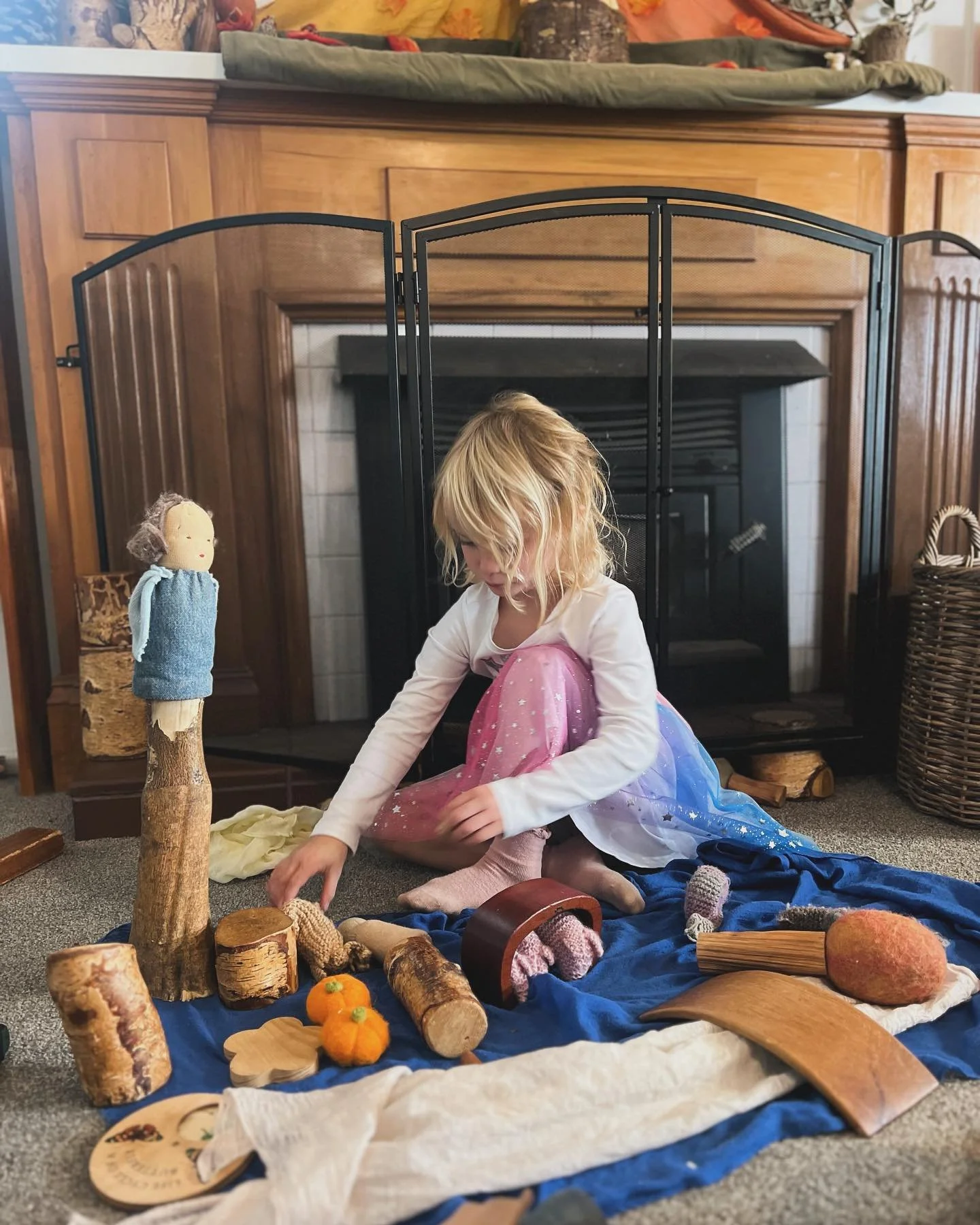 A young girl with blonde hair in pink and blue pajamas is sitting on a blue blanket on the carpet, playing with various wooden and fabric objects, including logs, a small pumpkin, and a doll, in front of a fireplace with a wooden mantle and a folding