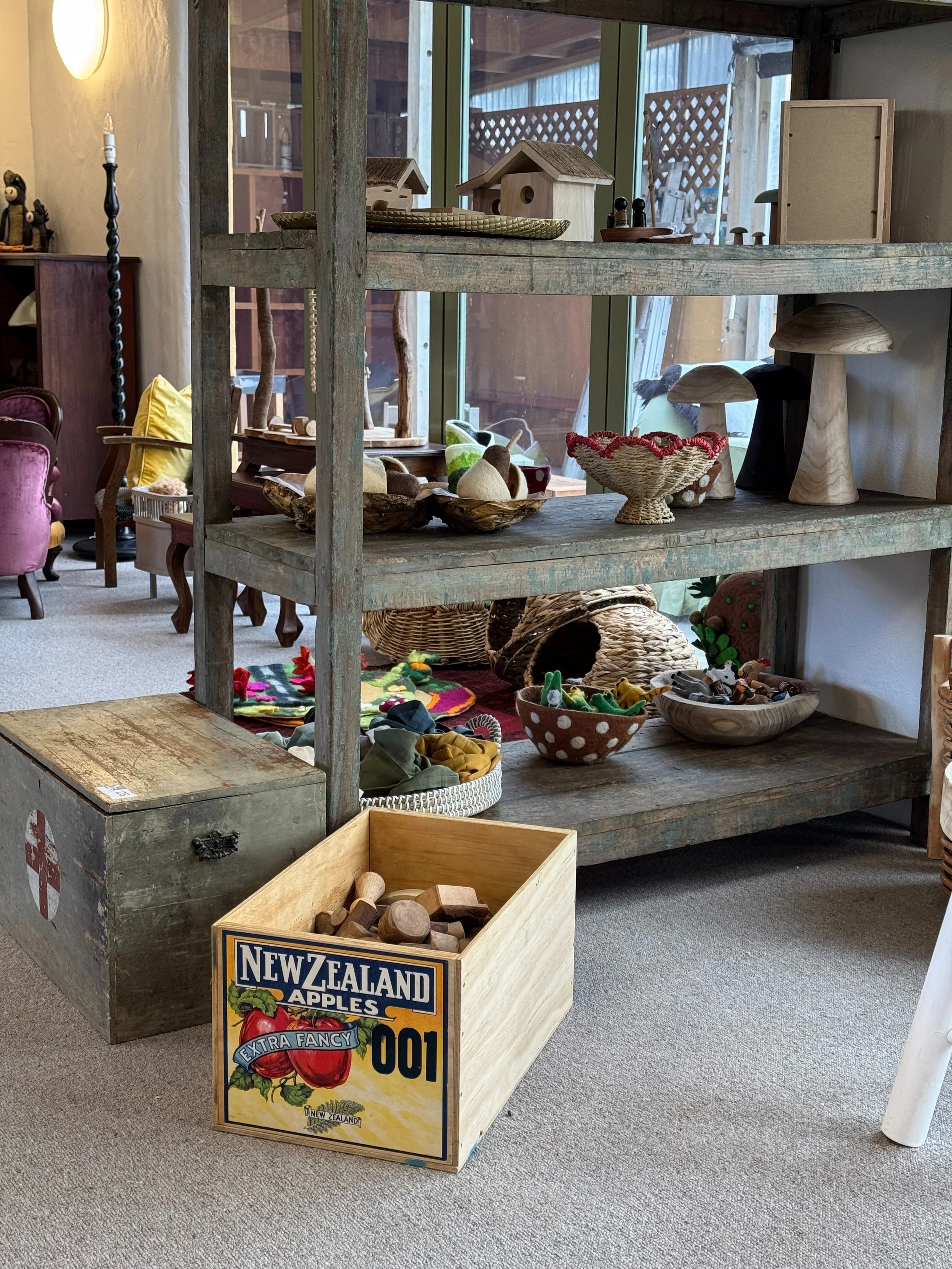 A wooden shelving unit displaying various small decorative and craft items, with a window in the background. A cardboard box labeled 'New Zealand Apples' sits on the floor in front of the shelf.