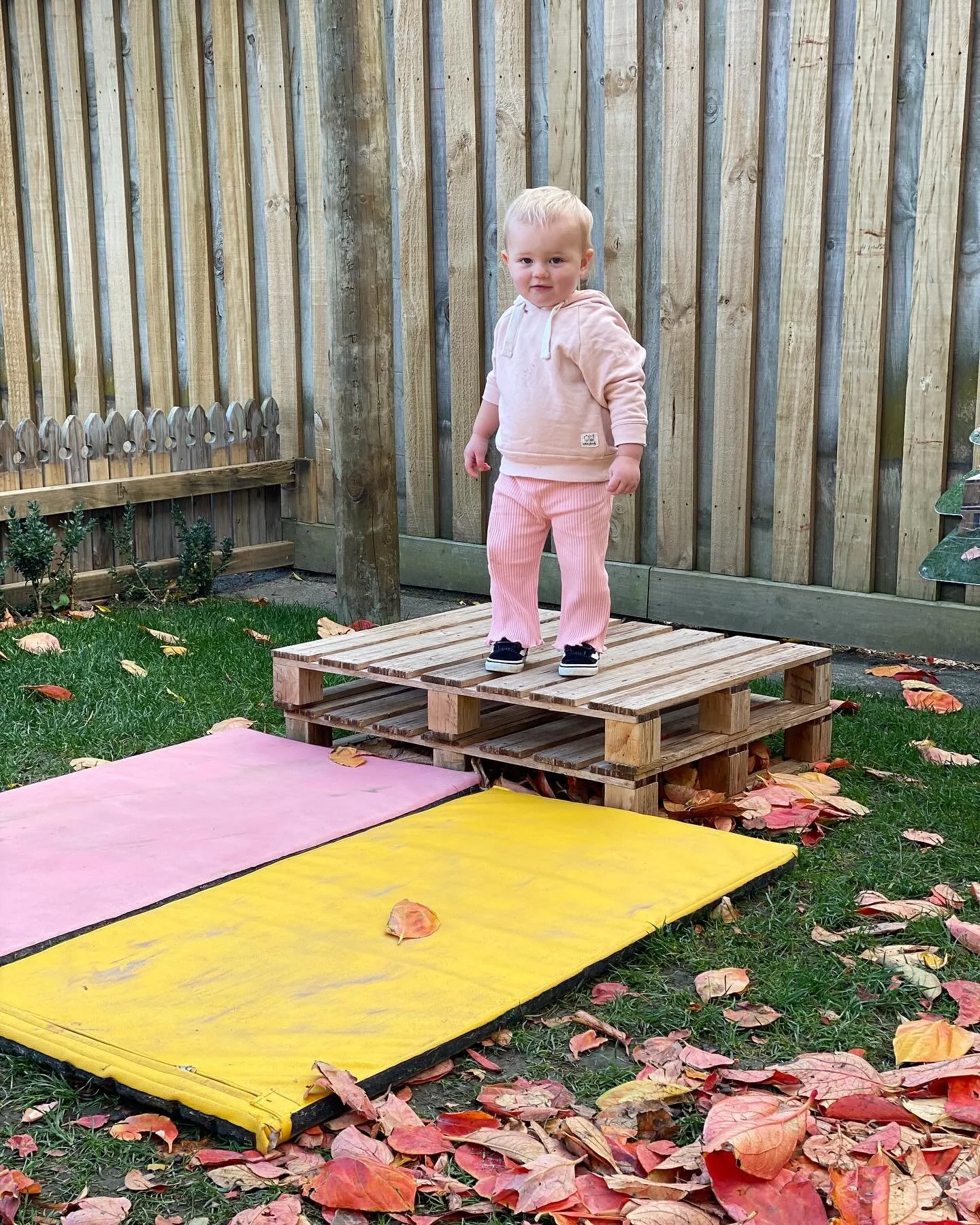 Child standing on a wooden pallet platform in a backyard with autumn leaves on the grass and colored mats on the ground.
