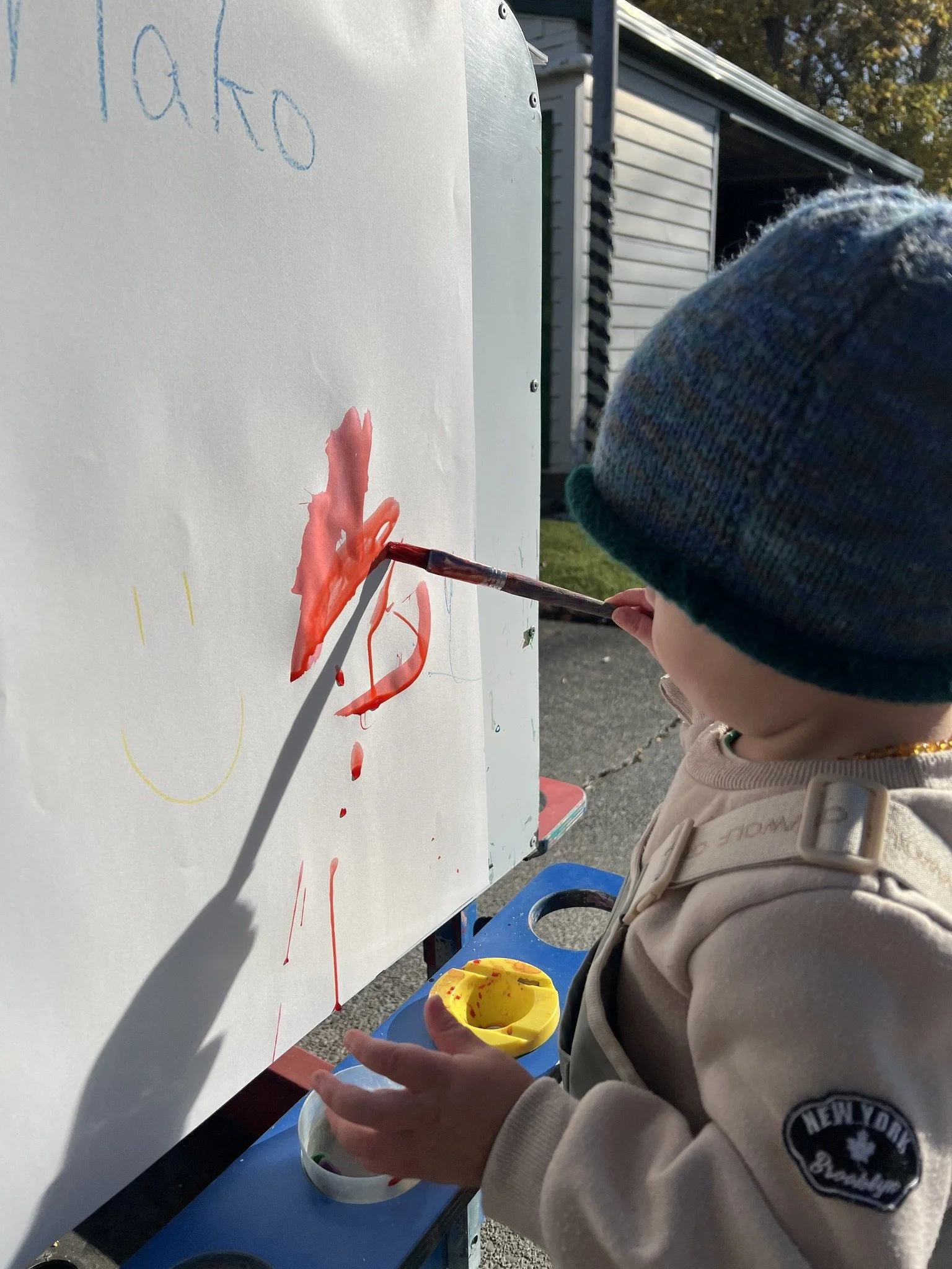 A young child, wearing a beige jacket with a patch that reads 'New York', a gold chain, and a blue knit hat, is painting with red paint on a large white surface outdoors. The child holds a paintbrush and is dipping it into a yellow paint container, w