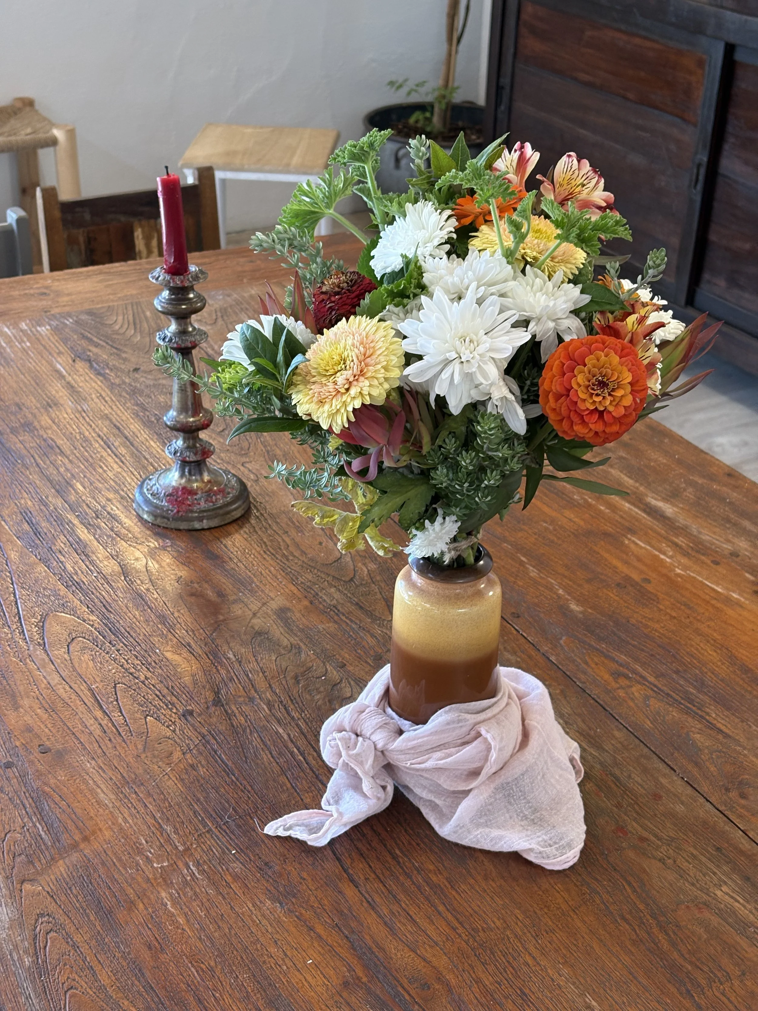A bouquet of colorful flowers in a brown glass vase on a wooden table, accompanied by a red candle in a silver holder.