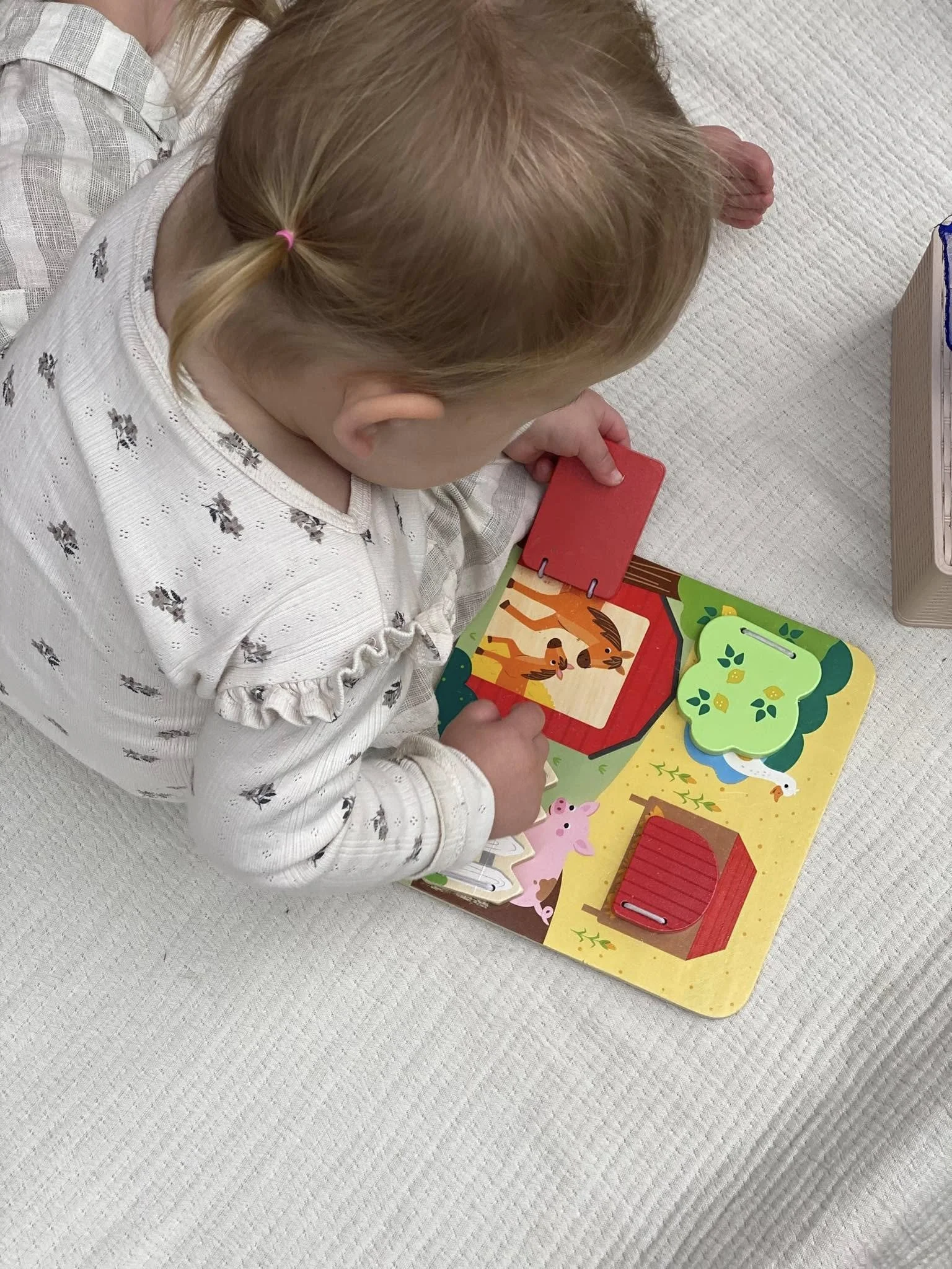 A young girl with blonde hair, wearing a white shirt, is playing with a colorful farm-themed wooden puzzle on a white textured surface.